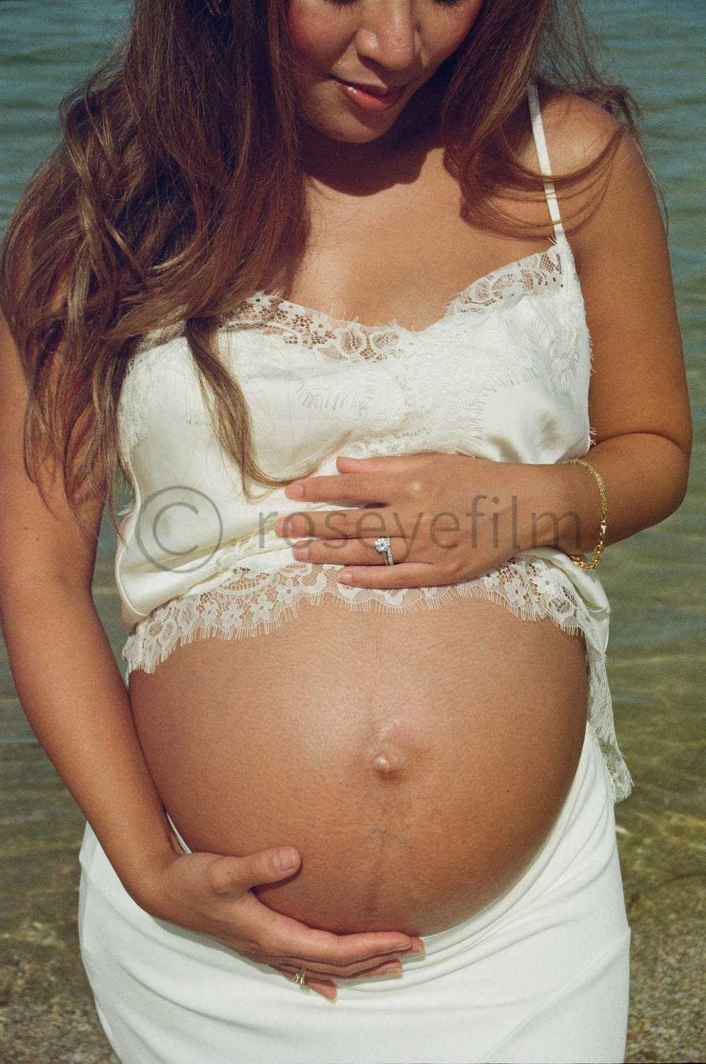 A pregnant woman stands by the water at the beach, wearing a white lace-trimmed top and white skirt, with one hand on her belly and the other on her chest.