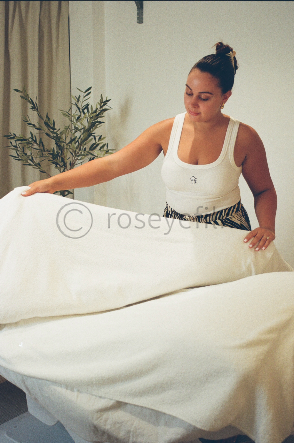 A woman in a white tank top and animal print skirt folds a white blanket or sheet on a bed in a room with beige curtains and a potted plant.