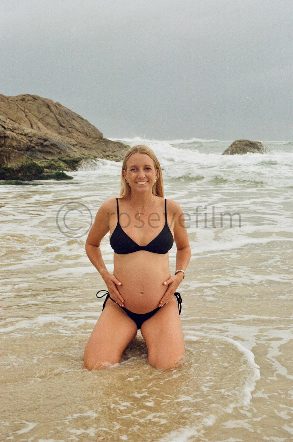 A pregnant woman in a black bikini kneeling in the ocean waves at a beach, smiling at the camera with rocks and cloudy sky in the background.