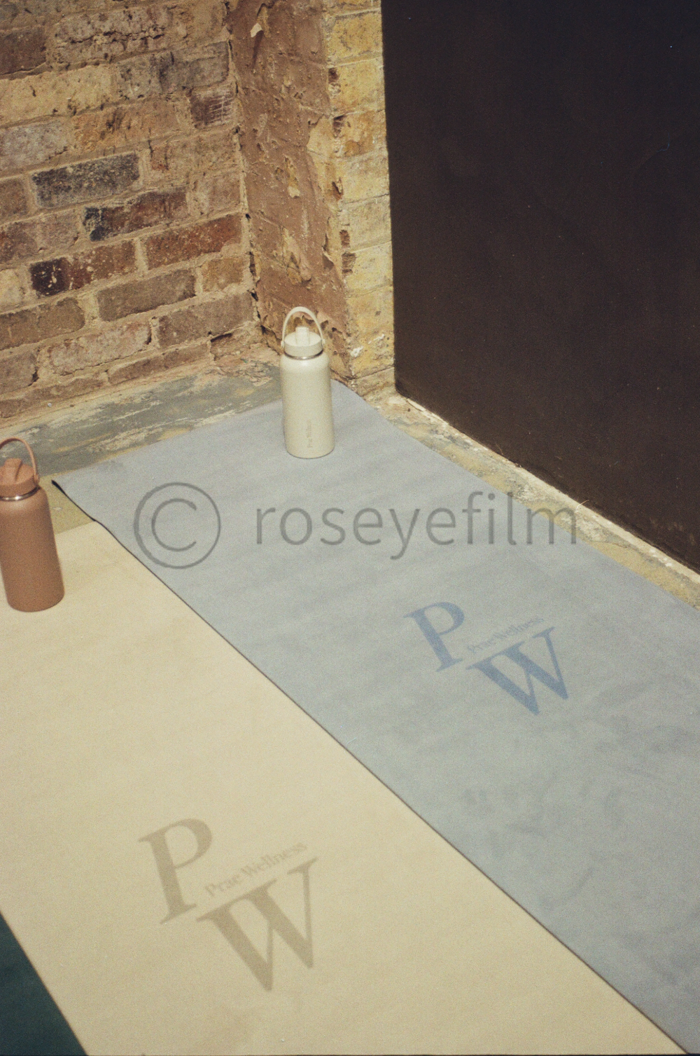 Two yoga mats, one beige and one light blue, with the P W logo printed on them, placed on the floor against a brick and concrete wall. Two water bottles, one beige and one white, are positioned on the mats.