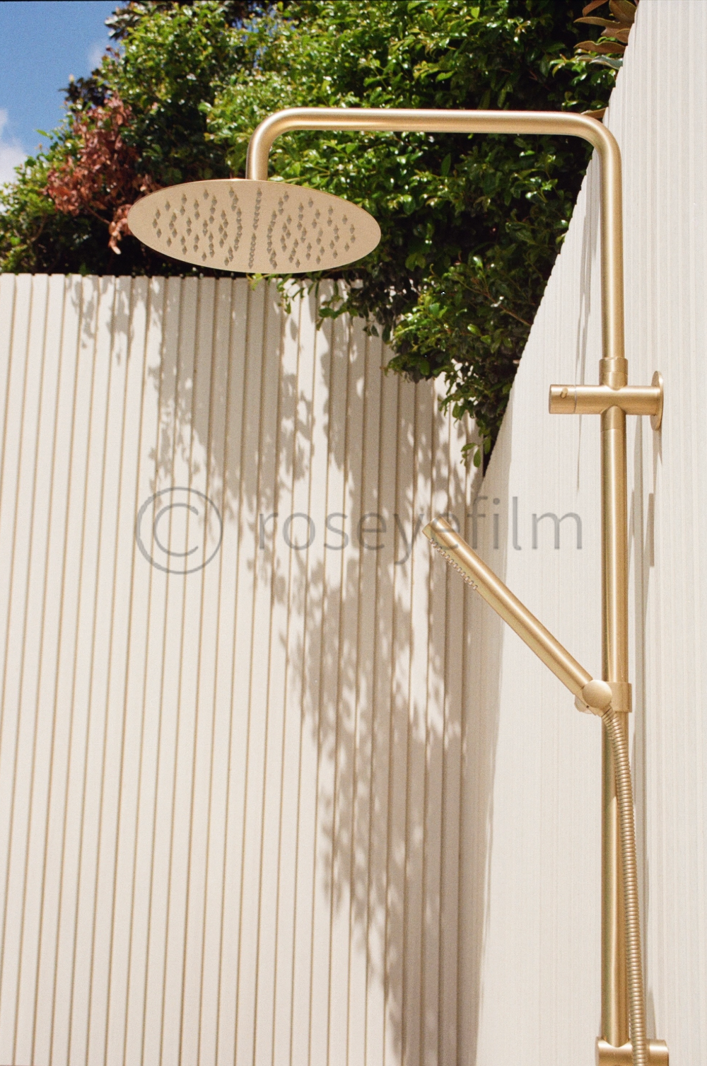 Outdoor modern shower with a gold finish, mounted on a white wall, with green foliage and a partly cloudy sky in the background.