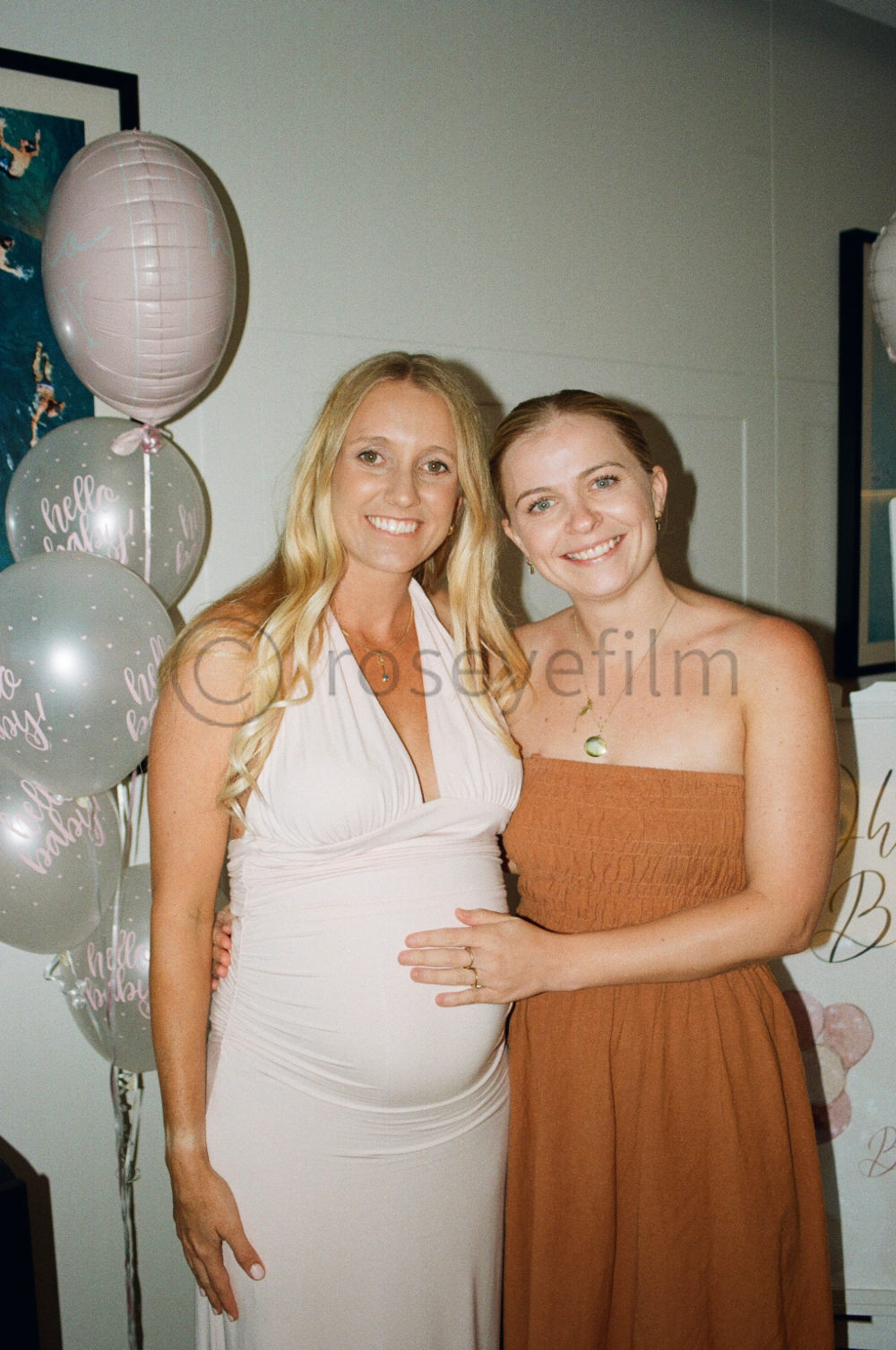 Two women standing close together at a celebration, one pregnant, smiling, with balloons in the background.