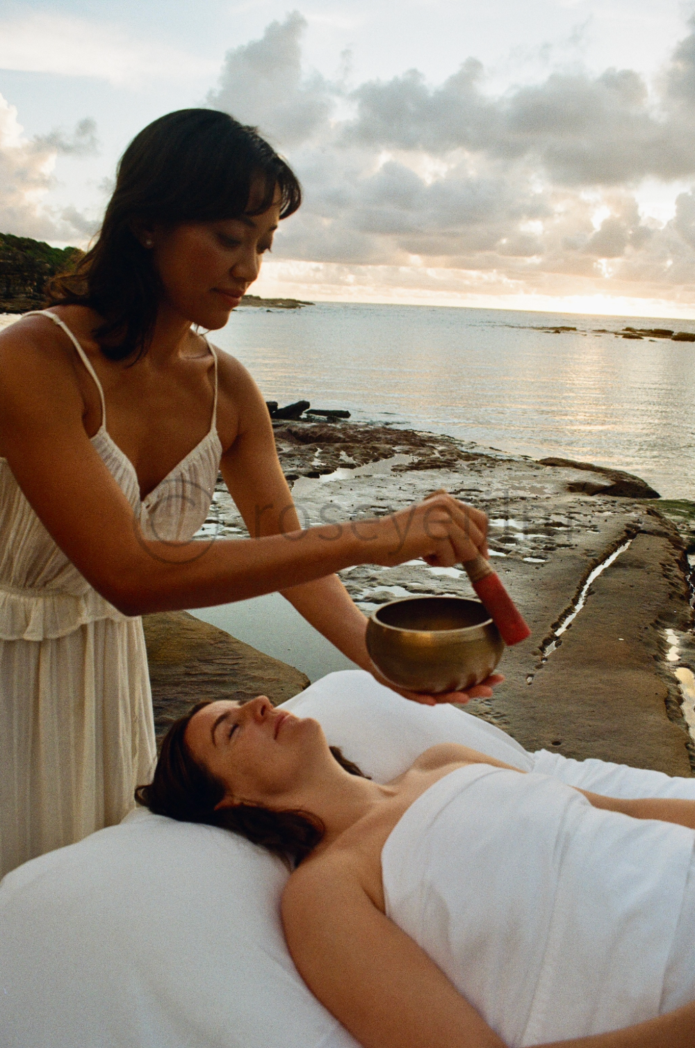 A woman in a white dress performs a sound bath with a singing bowl and mallet outdoors by the water at sunset, while another woman lies on a bed with her eyes closed.