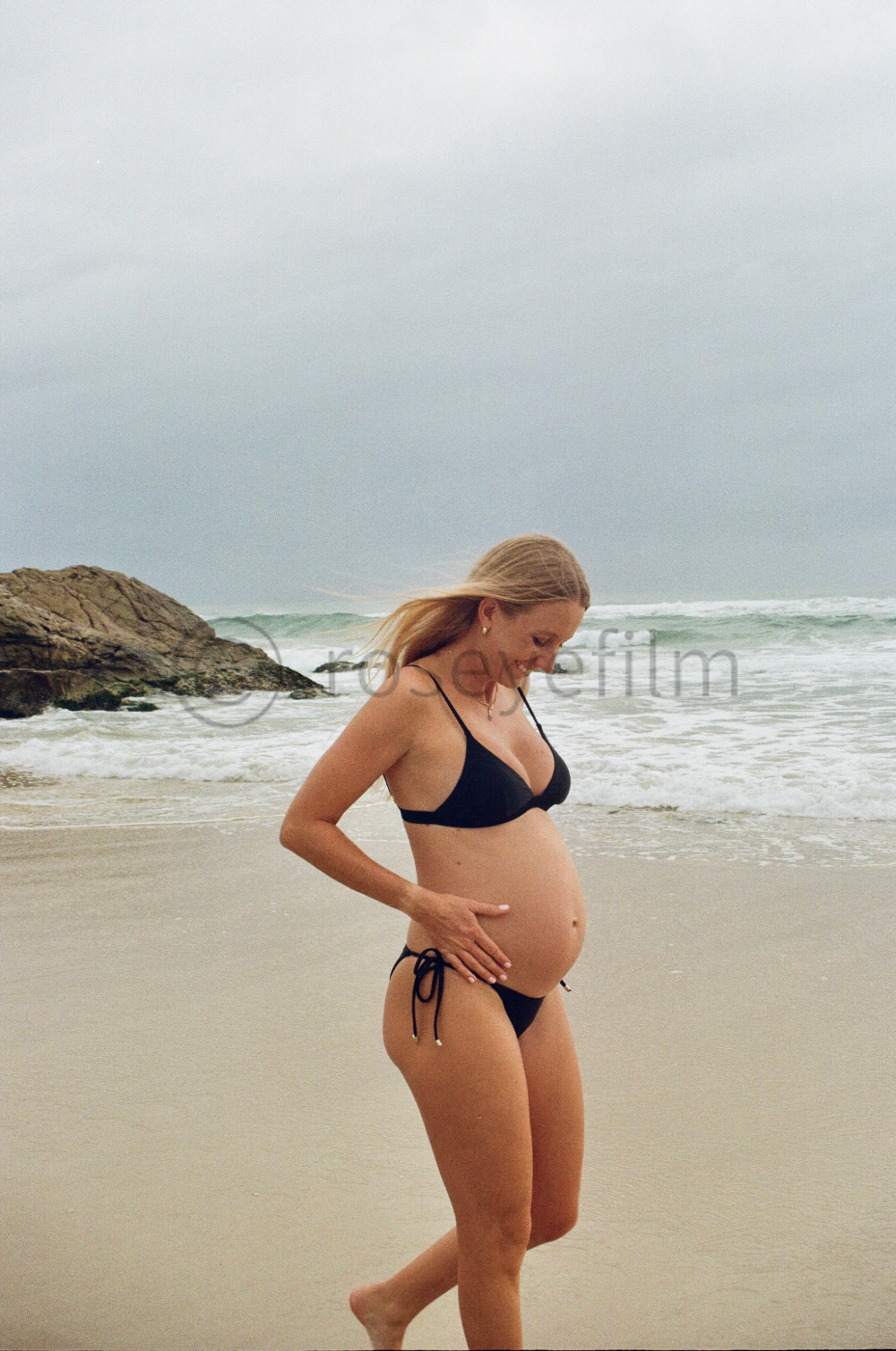Pregnant woman in a black bikini walking along the beach with rocks and ocean waves in the background.
