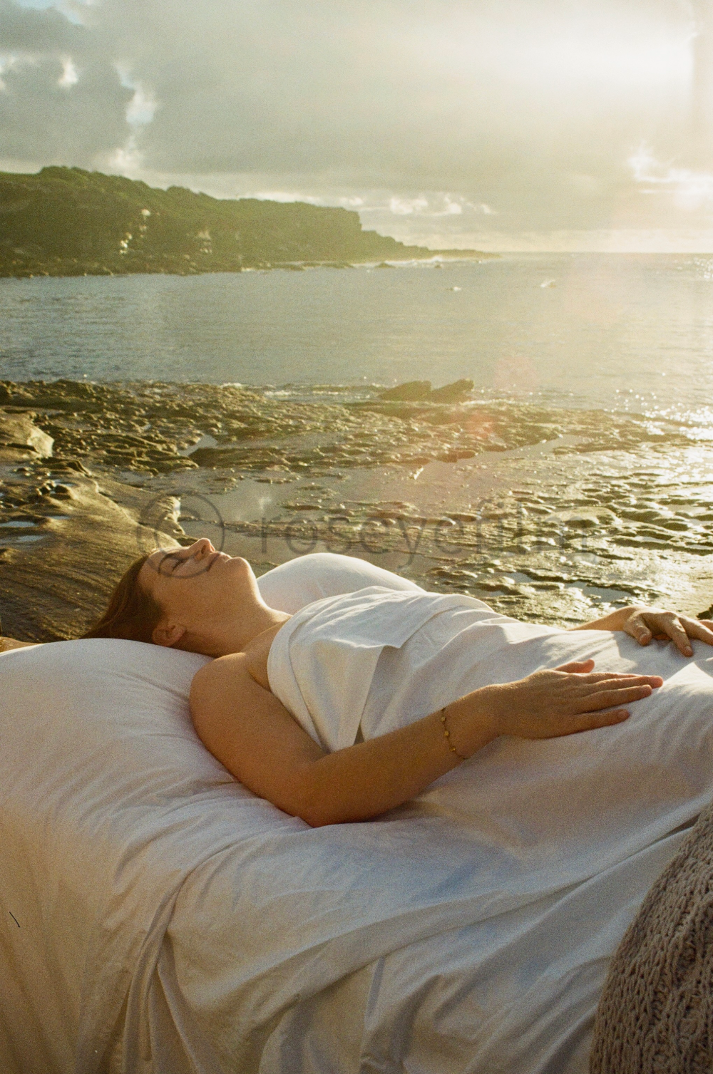 A woman in a white dress and glasses lying on a bed outdoors near a rocky beach, with water and hills in the background during sunset.