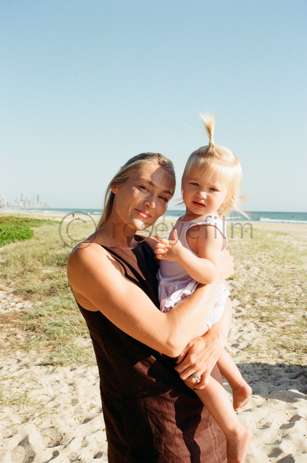 A woman holding a young girl on a beach with sand and ocean, city skyline in the distance, under clear blue sky.