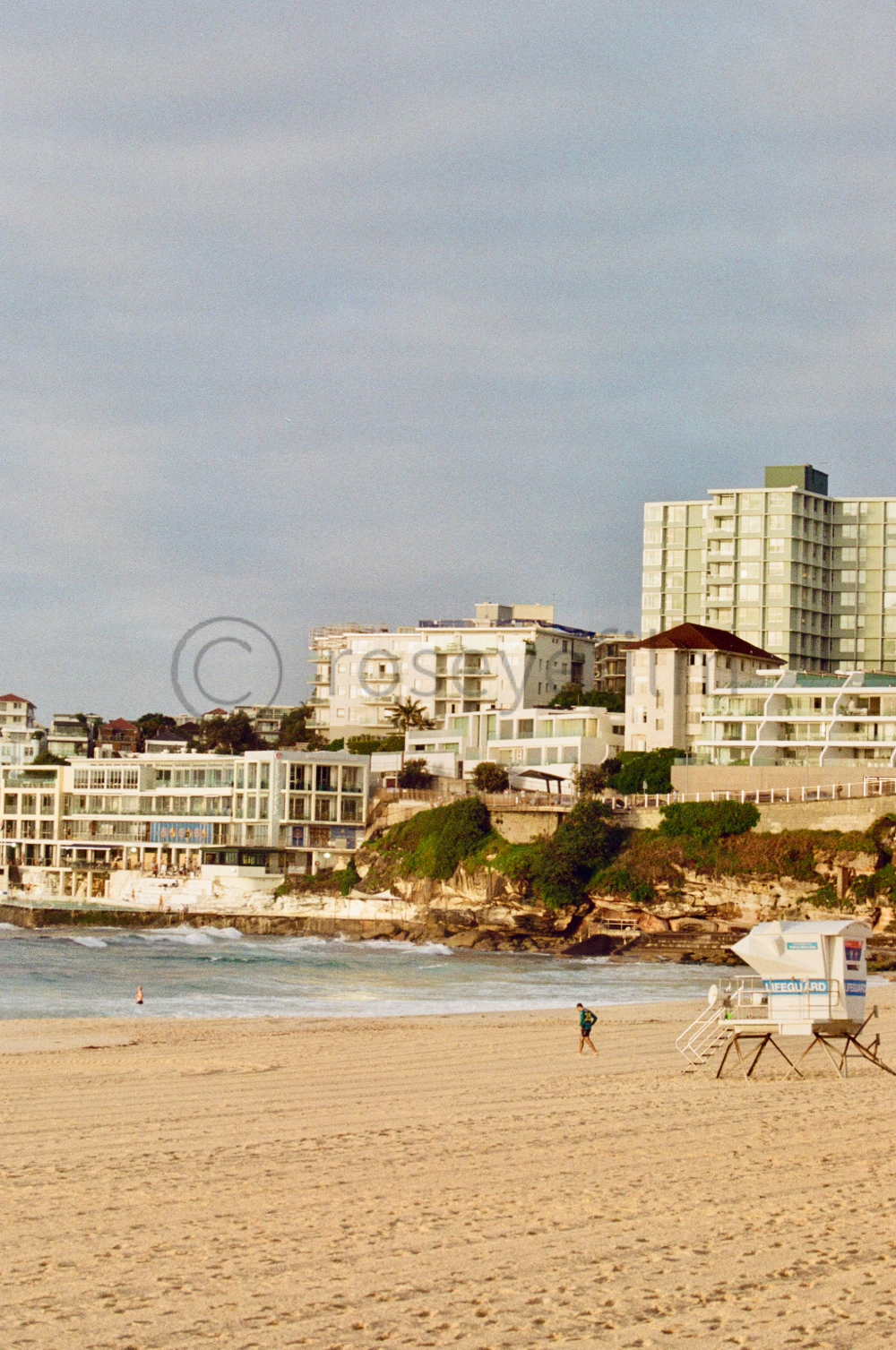 Sunlit beach with a lifeguard tower and two people walking along the shoreline. In the background, there are modern buildings on a hillside overlooking the beach.