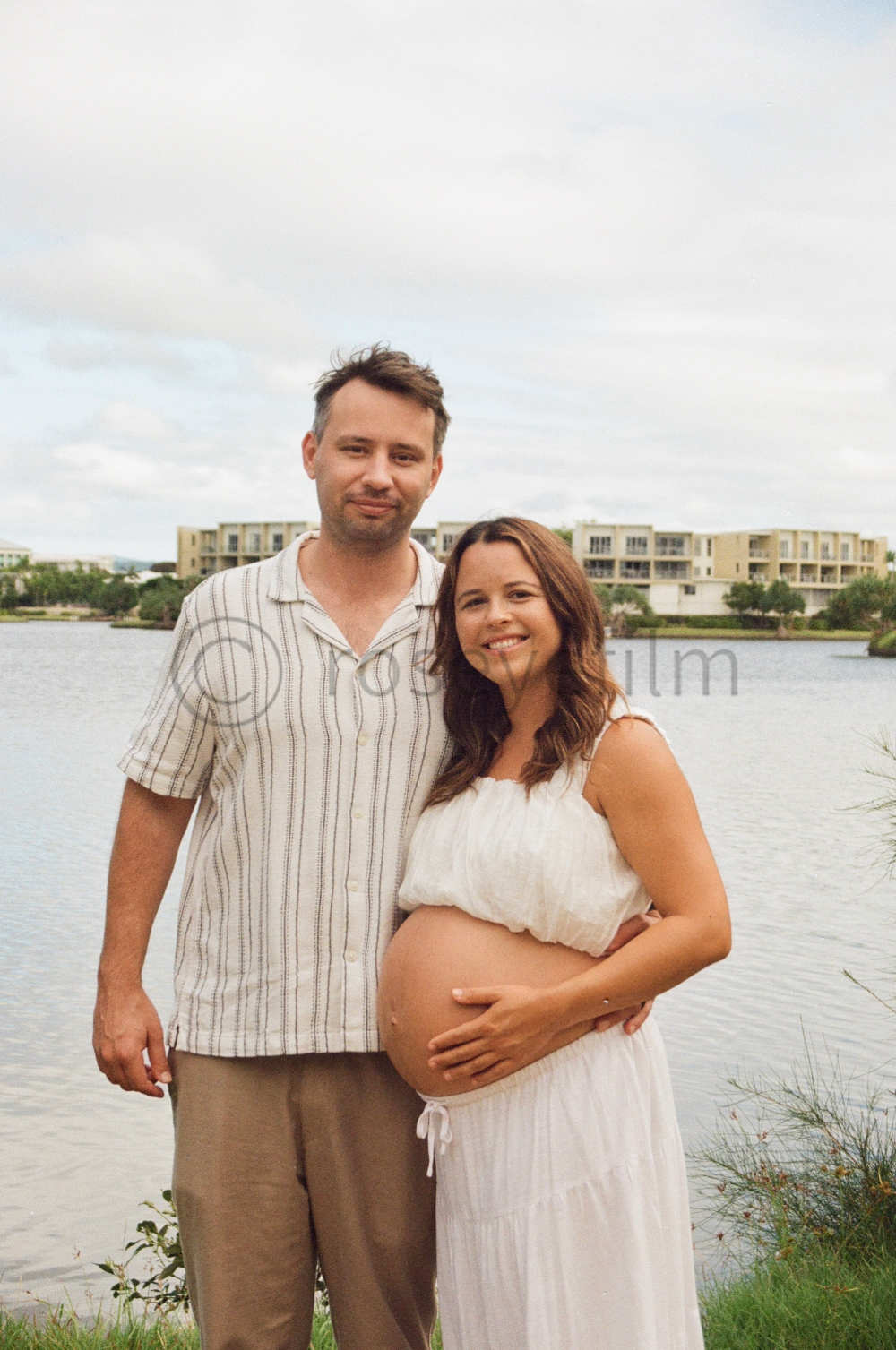 A couple standing outdoors by a body of water. The woman is pregnant, wearing a white dress, and smiling while holding her belly. The man is wearing a striped shirt and beige pants, standing beside her with a slight smile. In the background, there ar