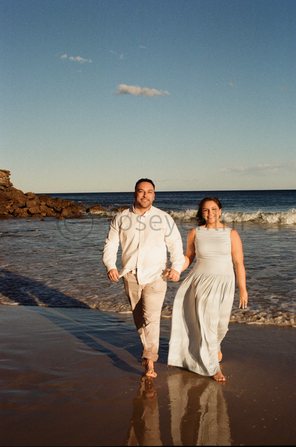A couple walking hand in hand on the beach during sunset, wearing light-colored clothing, with the ocean and sky in the background.