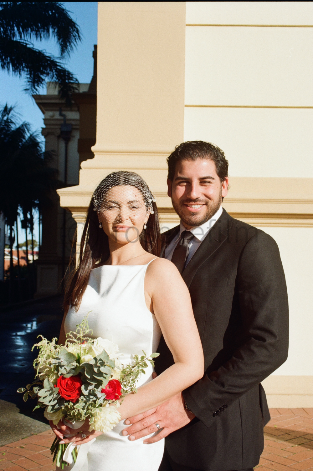 A bride and groom smiling outdoors on their wedding day, standing close together with the bride holding a bouquet of flowers, in front of a building with palm trees in the background.