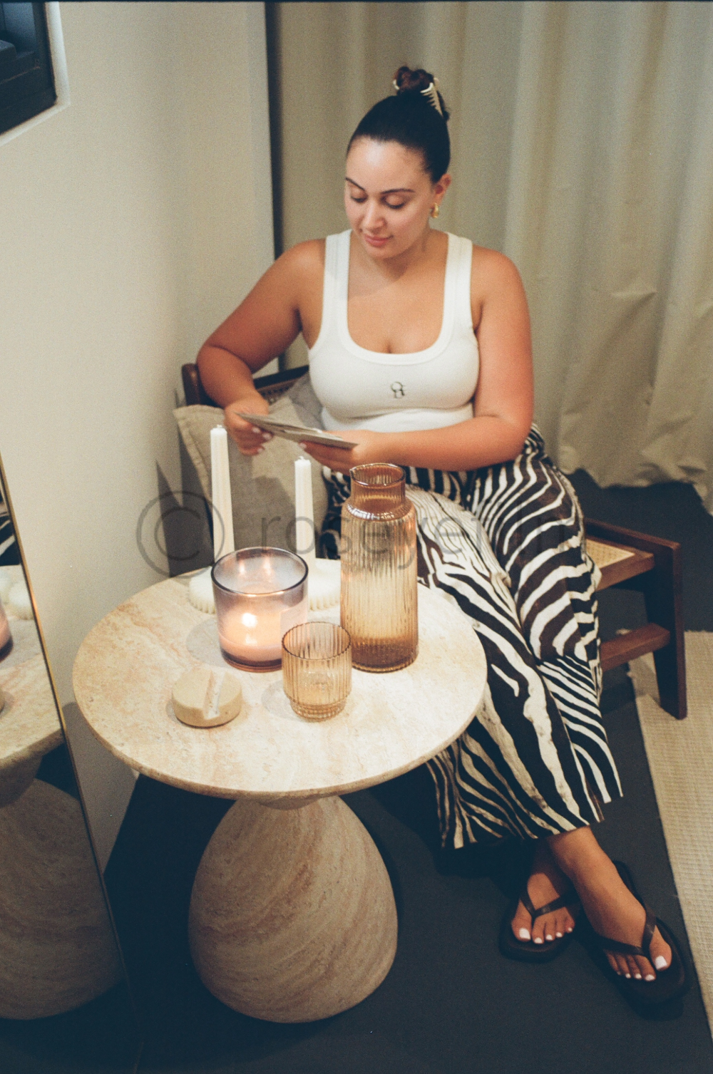 A woman sitting at a small round table decorated with candles, a vase, and glasses, looking at a photo or a piece of paper, dressed in a white tank top and zebra-print pants.