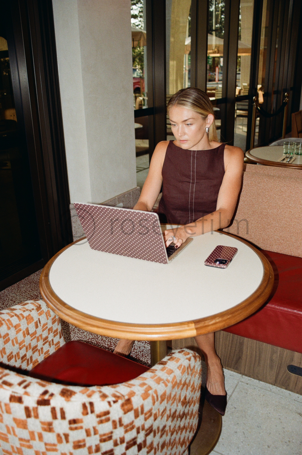 A woman in a sleeveless dark dress sitting at a round table inside a restaurant or cafe, working on a laptop with a polka-dot cover.
