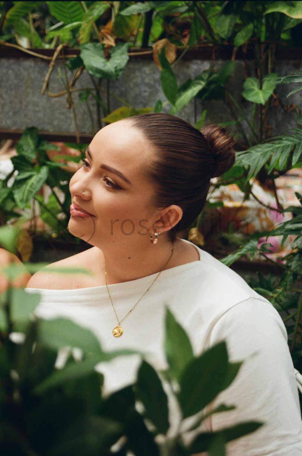 A woman with dark brown hair styled in a bun, wearing a white top, gold necklace, and small earrings, is surrounded by green plants and looking to her left with a slight smile.