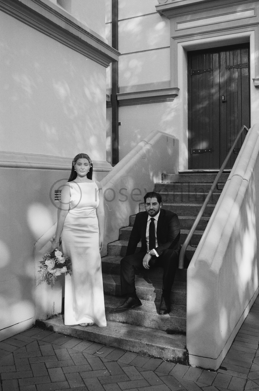 A woman in a long white dress holding a bouquet of flowers standing on stairs beside a seated man in a dark suit and tie, outside a building.