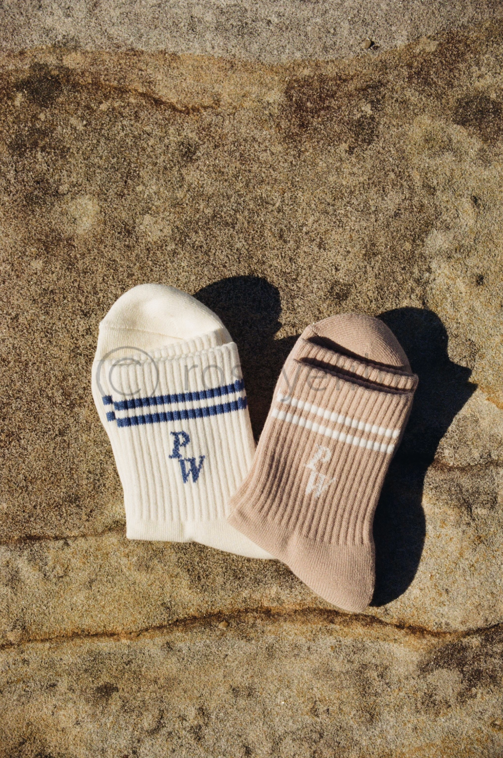 Pair of beige and white striped ankle socks with white embroidered initials on a sandy surface.
