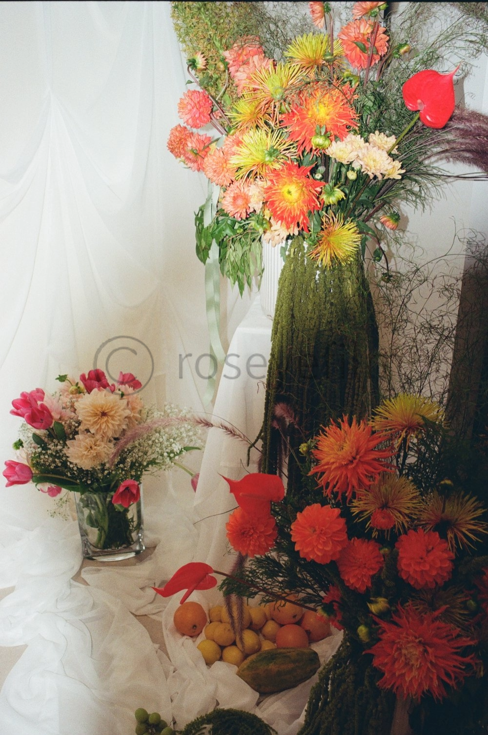 Colorful floral arrangement with orange and yellow flowers in a tall green moss-covered vase, accompanied by a smaller pink and white flower bouquet in a glass vase, set against a white draped background with white fabric and fruit on a white cloth.