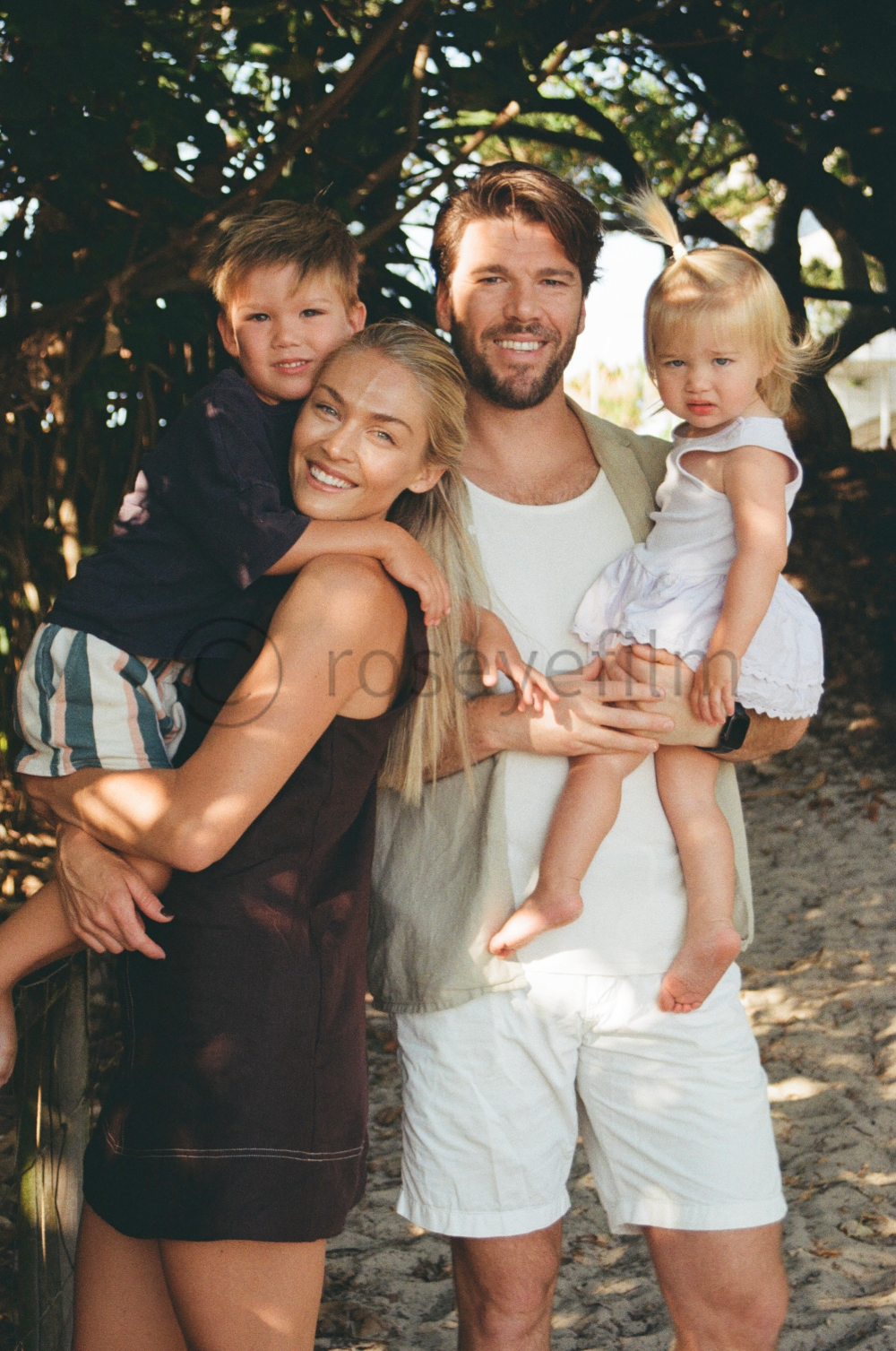A happy family of four outdoors with trees in the background. The mother, with long blonde hair, smiles as she carries a young boy on her back. The father, with dark hair and a beard, holds a young girl in a white dress. They are enjoying a sunny day