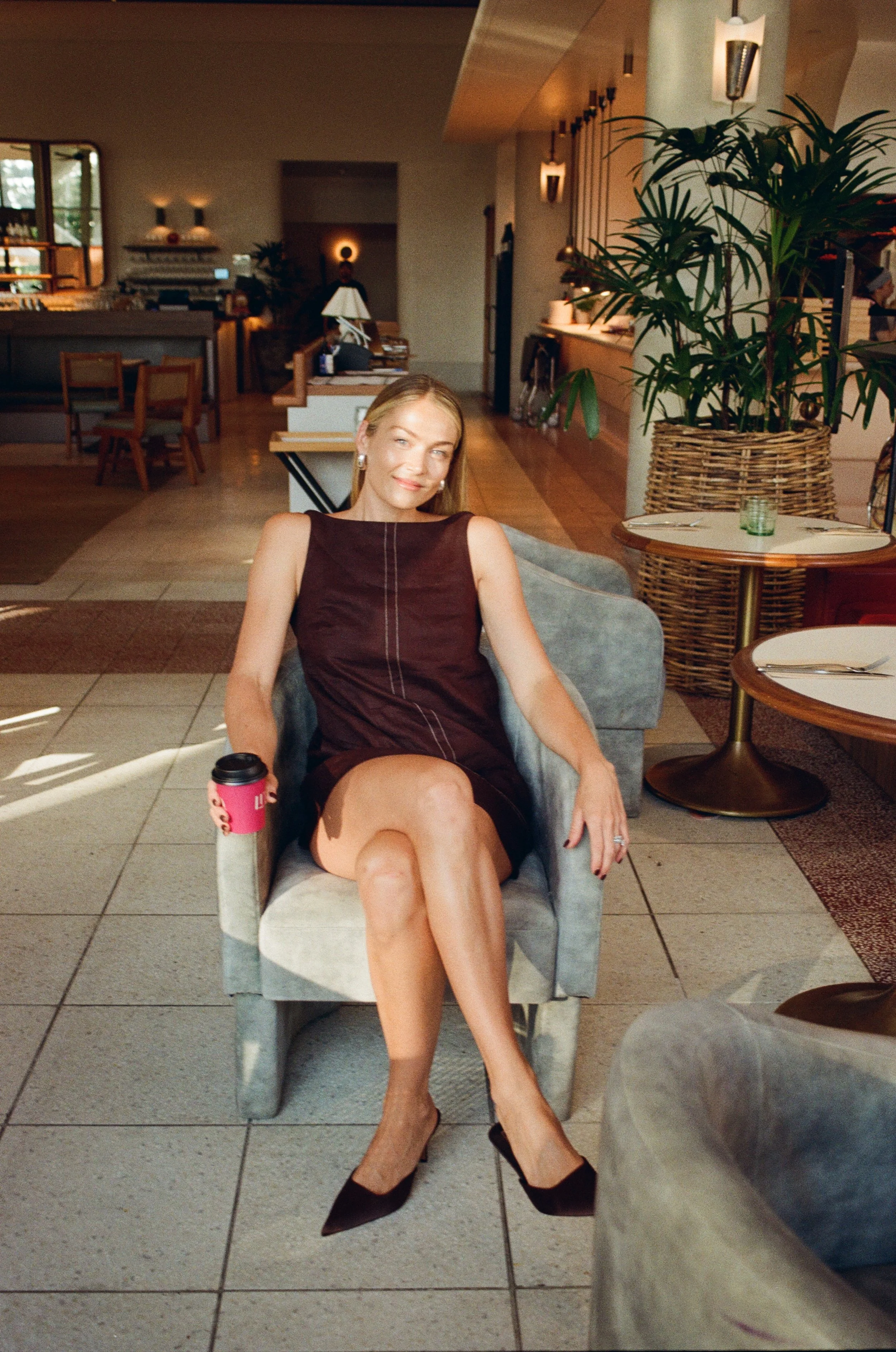 A woman with blonde hair, wearing a dark sleeveless dress and black heels, seated in a light-colored armchair in a restaurant or lounge with wooden floors, a potted plant, and tables set for dining.