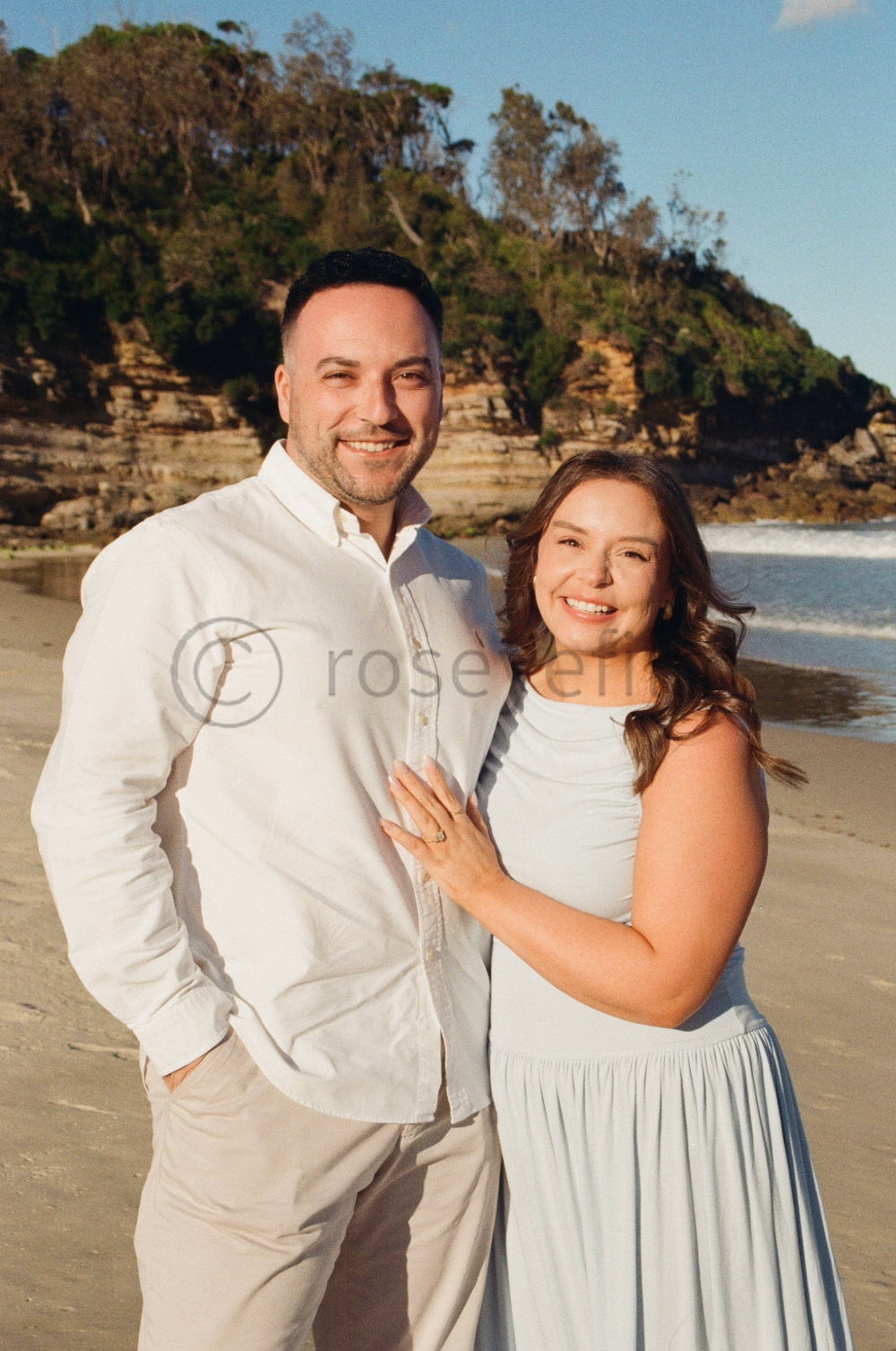 A smiling couple standing on a beach with a rocky hill and trees in the background during sunset.