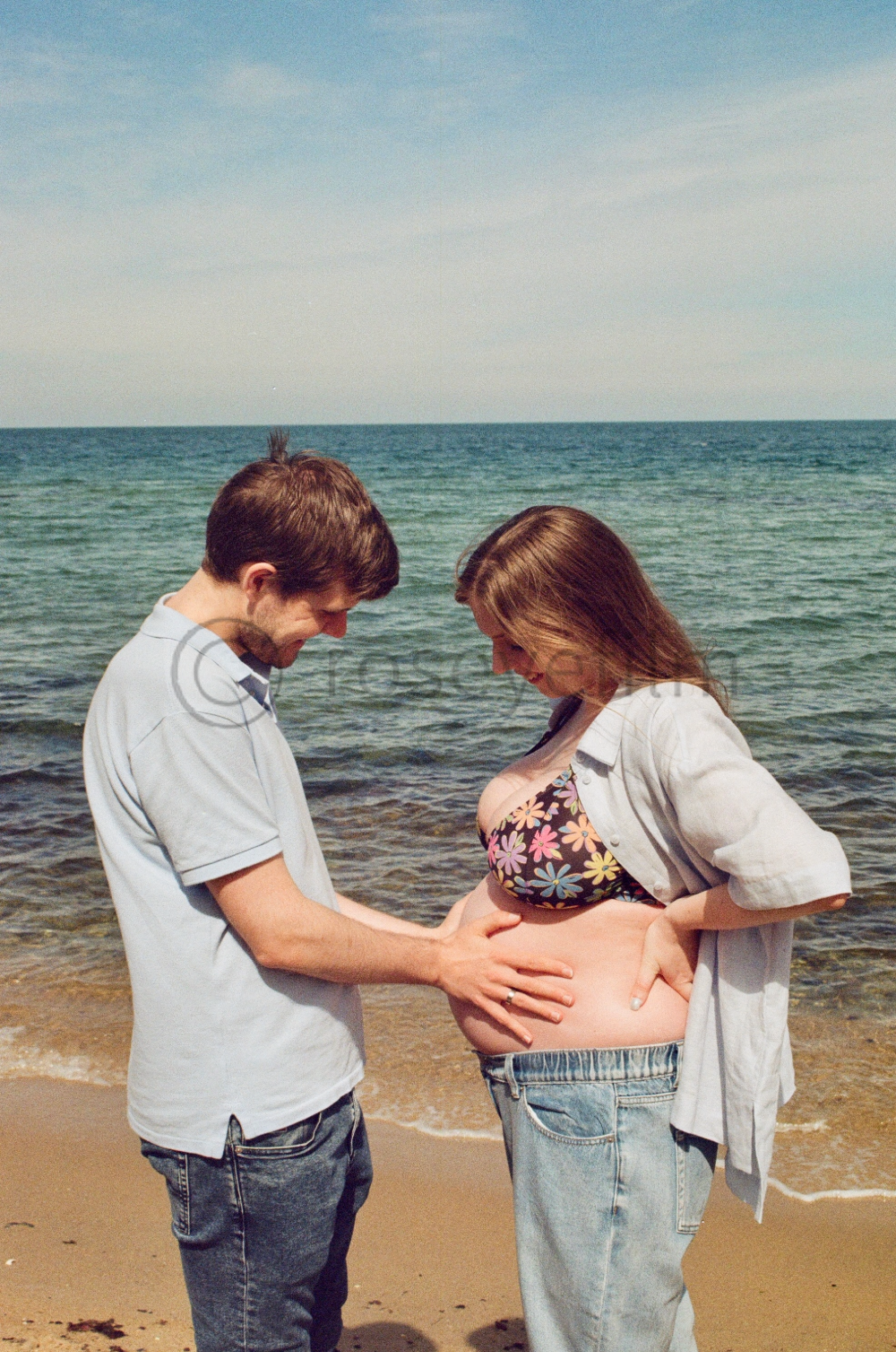 A couple stands on the beach with the ocean and sky in the background. The woman is pregnant and is wearing a floral bikini top and jeans. The man is touching her belly, and they are both smiling and looking down at it.