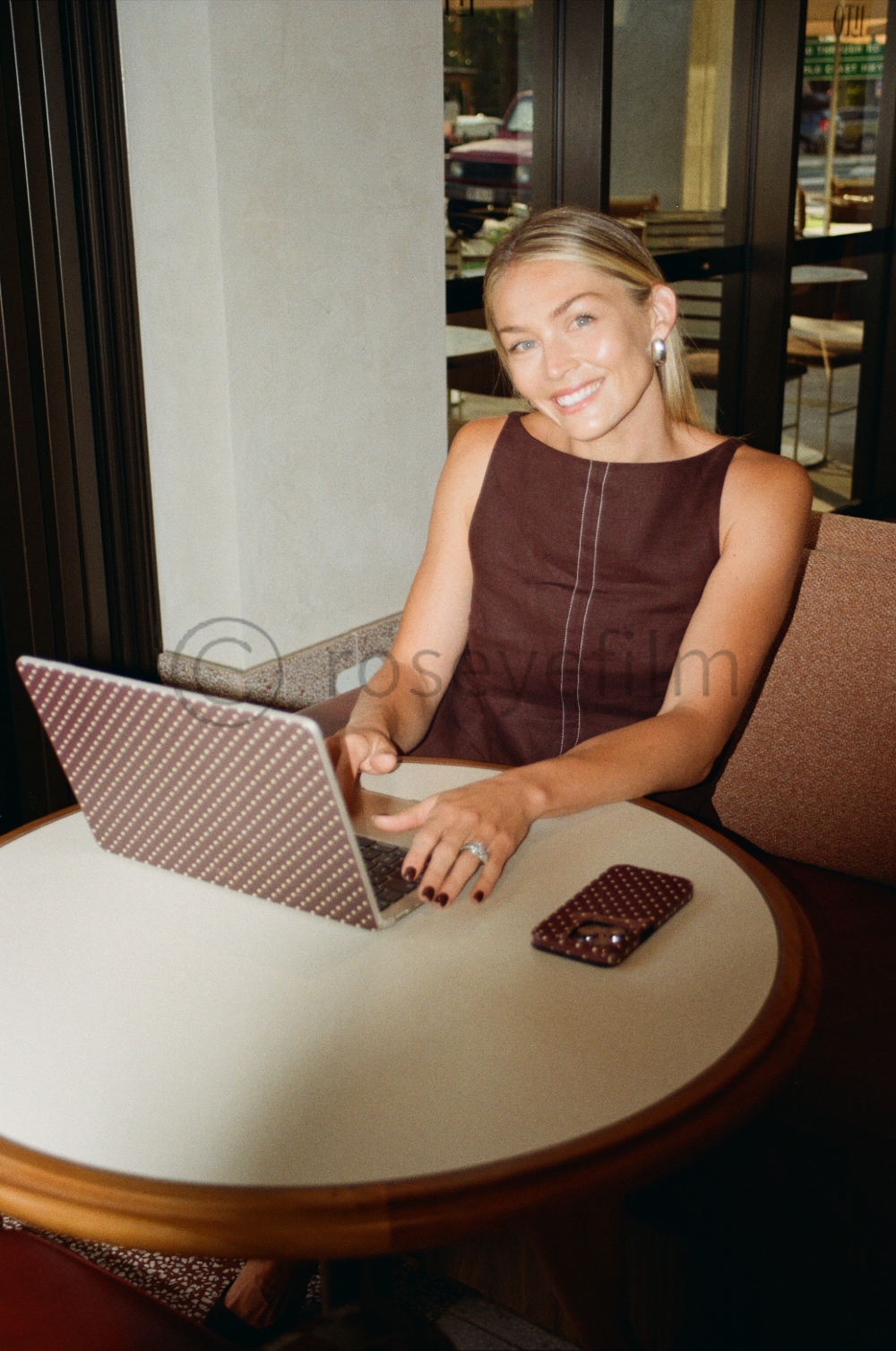 A smiling woman with blonde hair, wearing a sleeveless brown top, sitting at a round table in a cafe, using a laptop with a matching patterned cover, and a smartphone with a similar cover placed on the table.