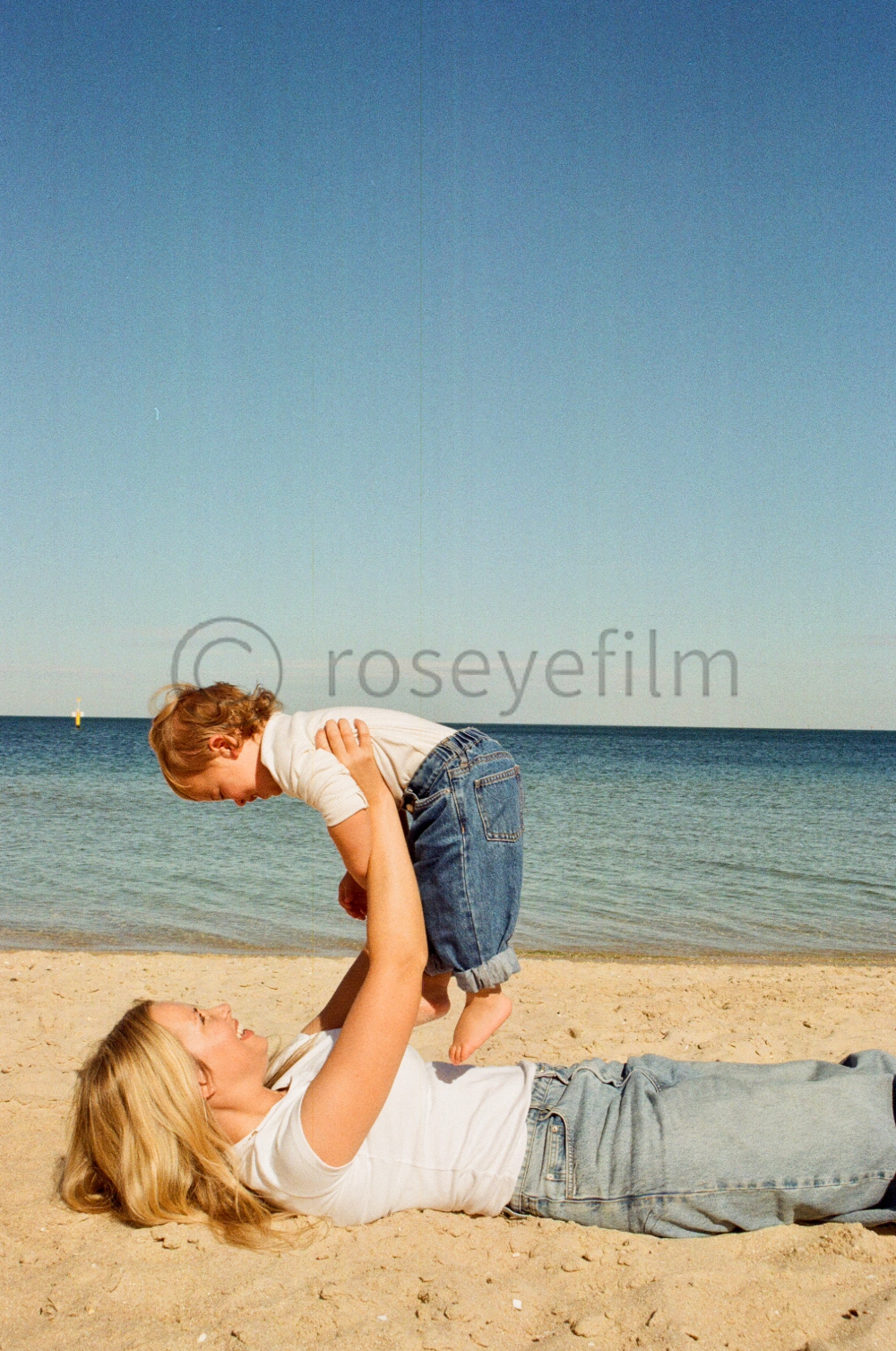 A woman lying on the beach, holding a young boy in the air, with the ocean and a clear sky in the background.