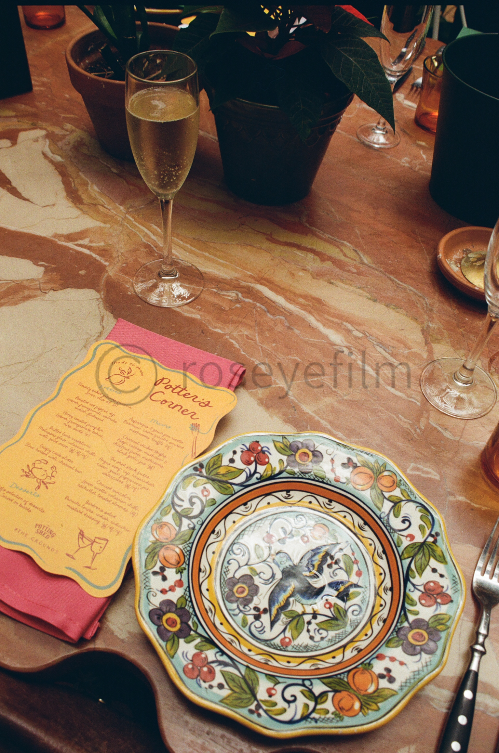 A dining table set with decorative plates, a menu, and glasses of sparkling wine. There are potted plants and various table items scattered around.