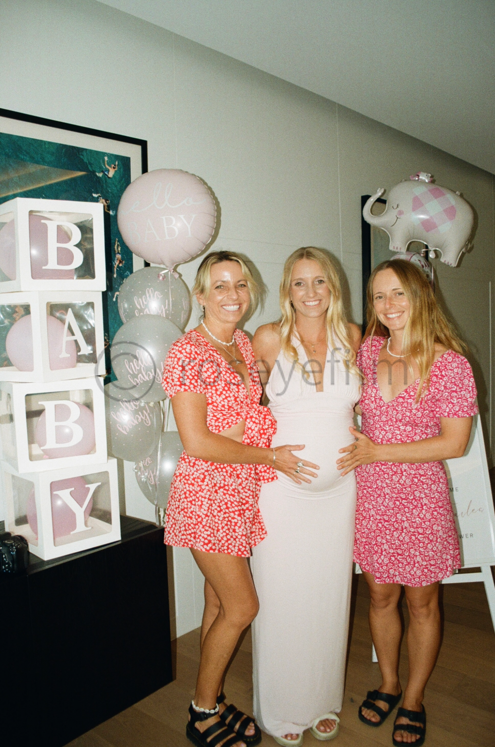 Three women celebrating a pregnancy at a baby shower, with balloons and decorative blocks spelling 'BABY' in the background.