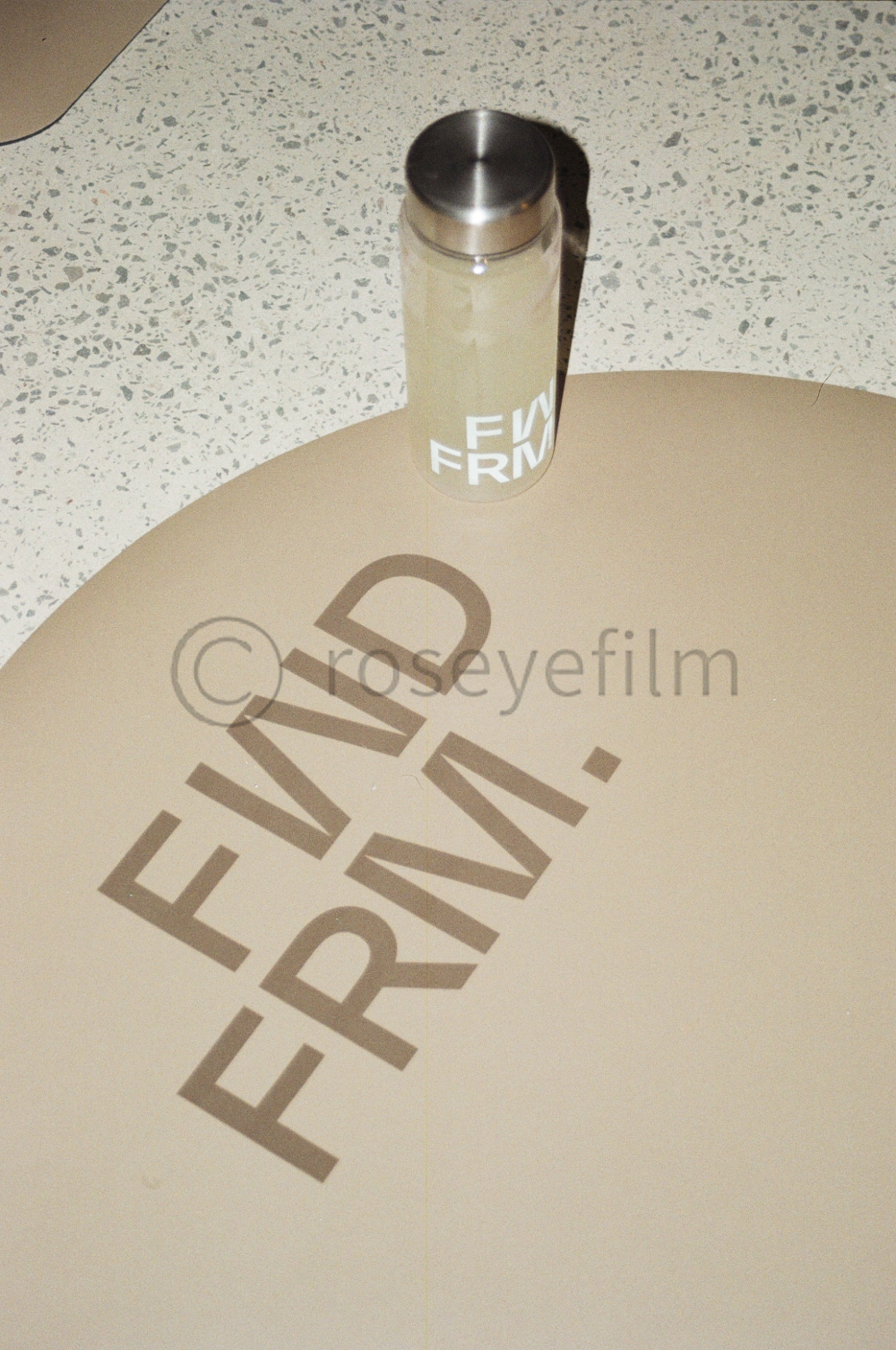 A beige table with the words 'EERING' and 'NDR' printed on it, and a beige tumbler with a stainless steel lid casting a shadow on the table.