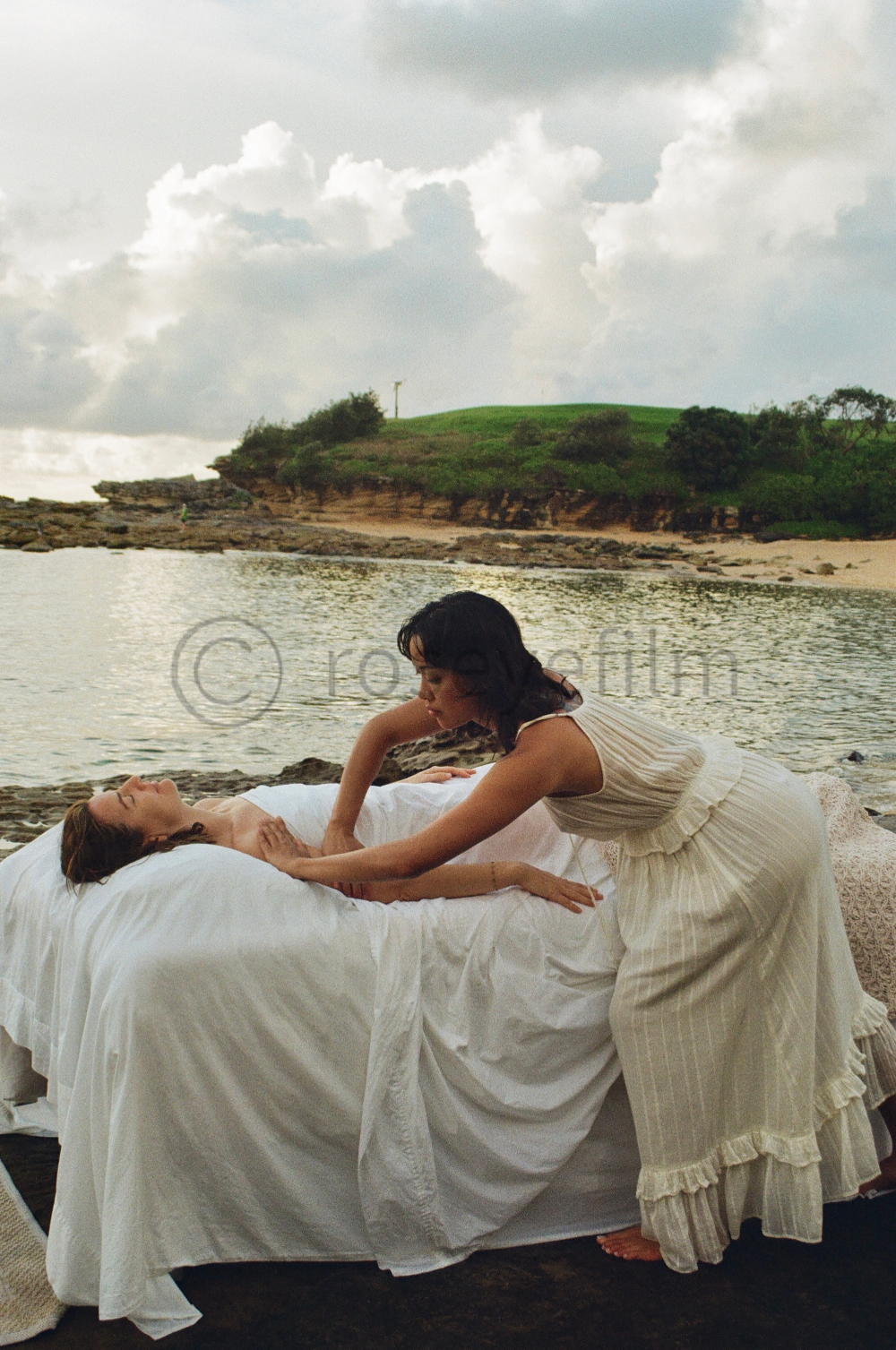Two women in white dresses are practicing reiki or massage therapy outdoors on a rocky beach with a green hill and cloudy sky in the background.