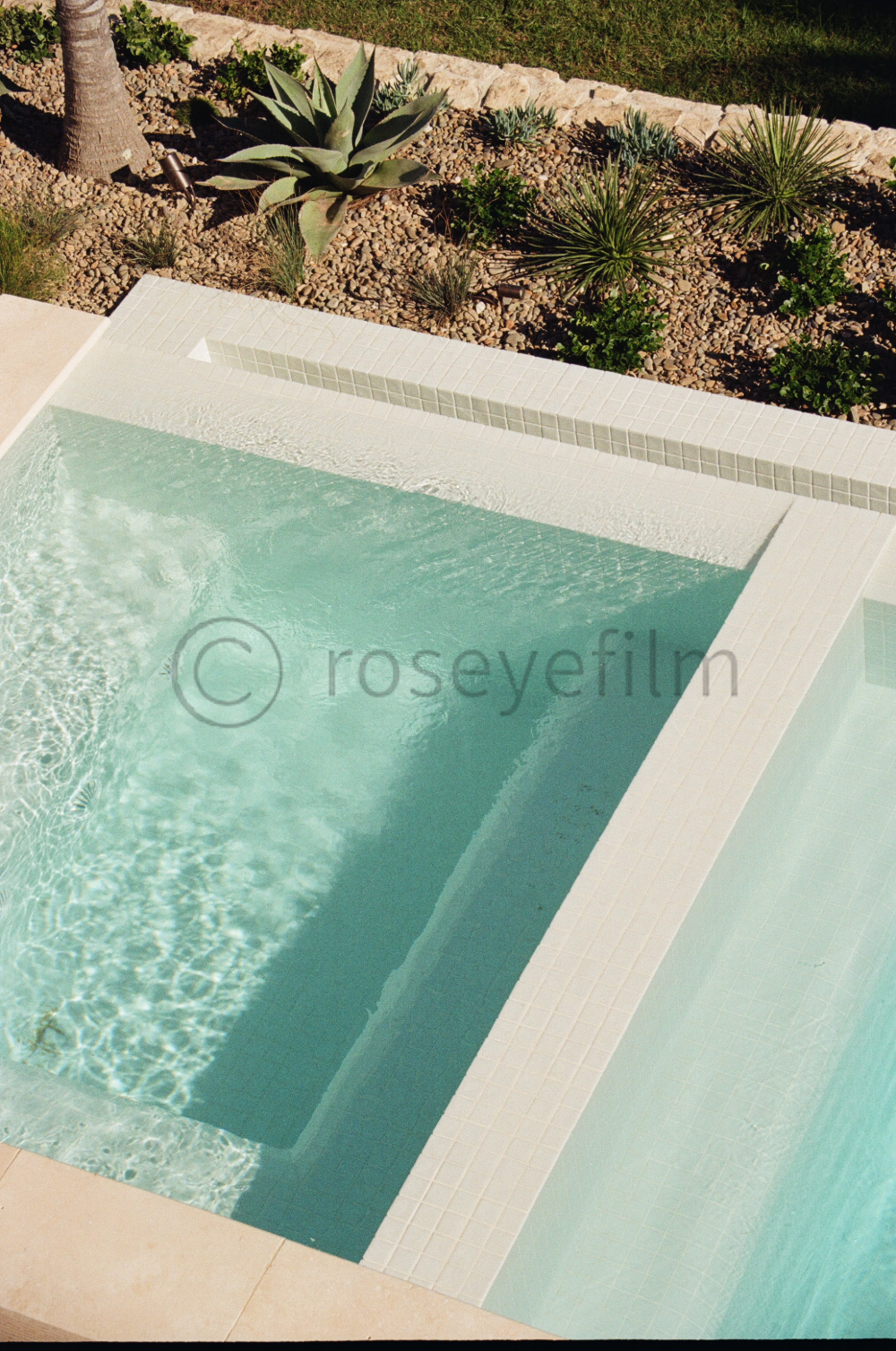 A swimming pool with clear water bordered by white tiles, with desert plants and rocks in the background.
