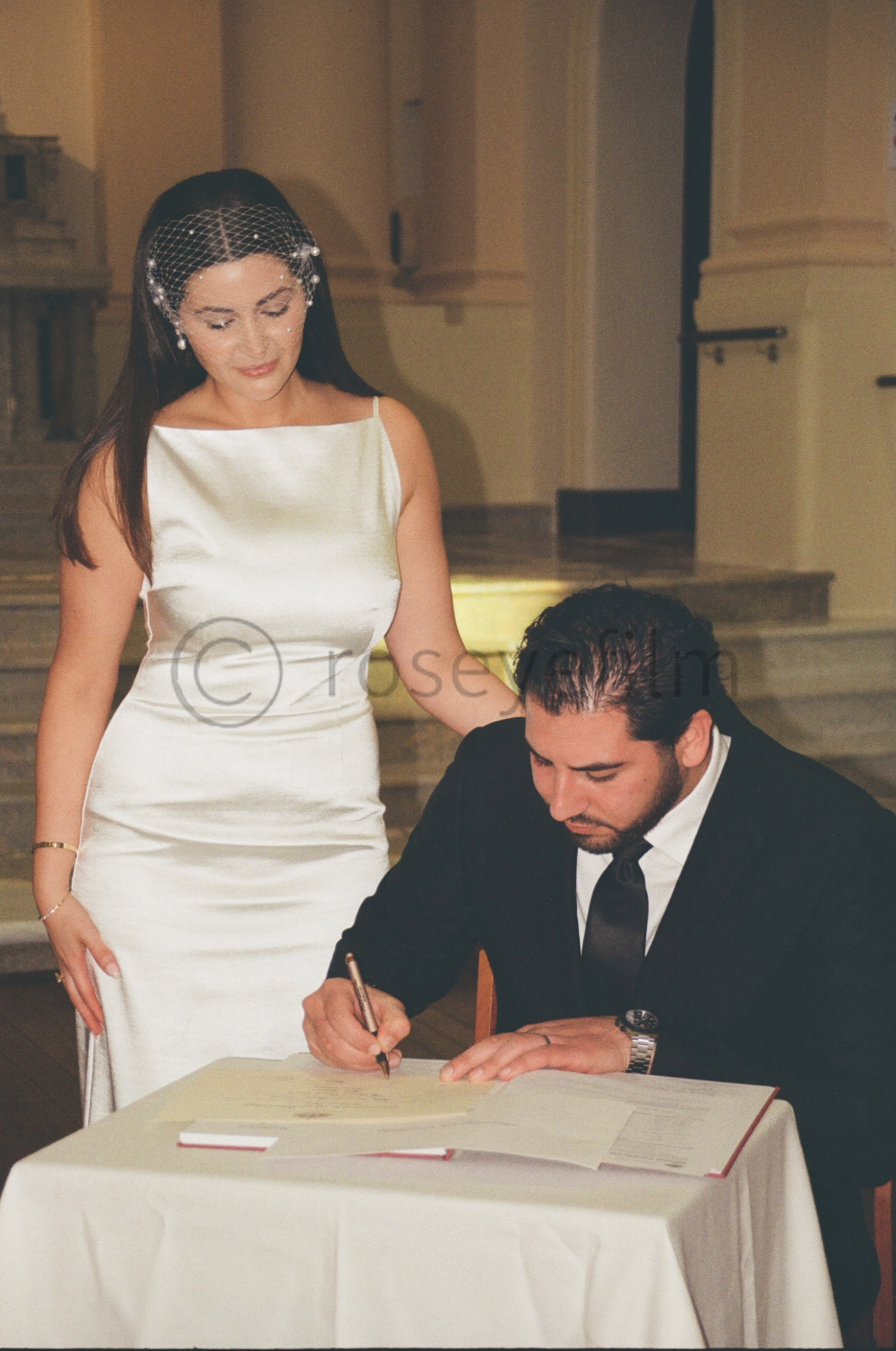 A woman in a white satin dress and a man in a black suit and tie are at a table, with the woman observing as the man signs a document, likely during a wedding ceremony.