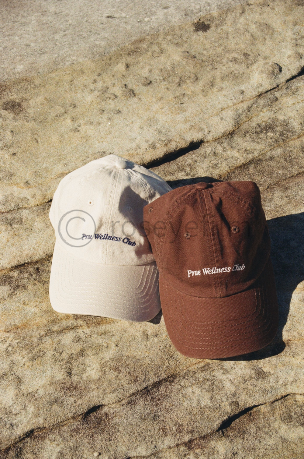 Two baseball caps, one white and one brown, with 'Prime Wellness Club' embroidered on the front, resting on a large, textured rock surface.