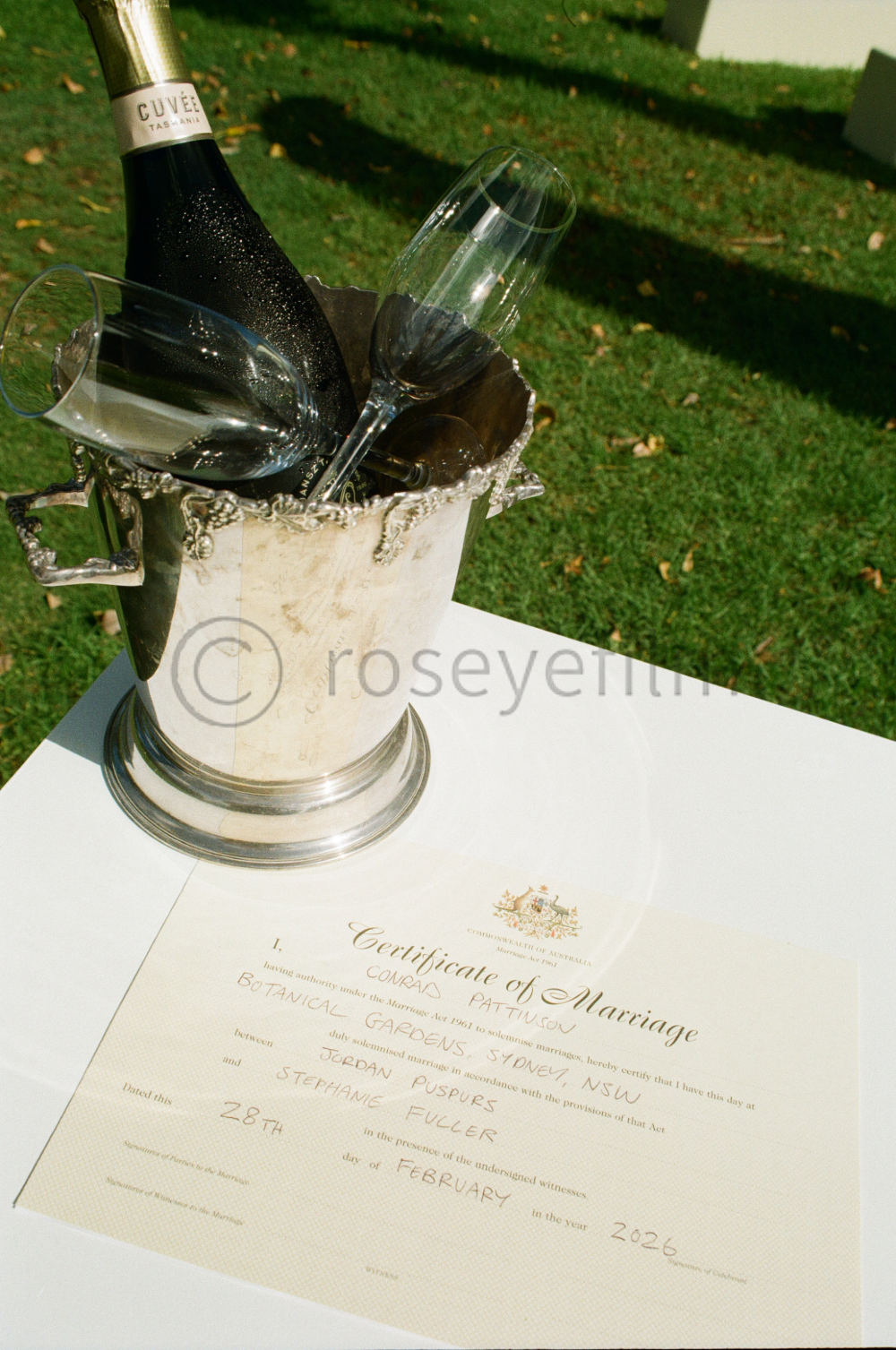 A wedding certificate of marriage placed on a white table with a silver ice bucket containing a bottle of sparkling wine and two champagne glasses, set outdoors with green grass in the background.