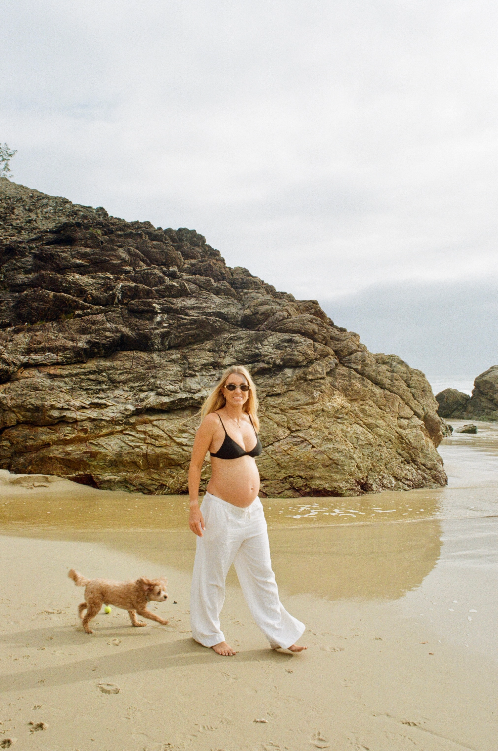 A pregnant woman wearing a black bikini top, white pants, and sunglasses walking on the beach with her small brown dog during daytime. Large rocks and the ocean are in the background.