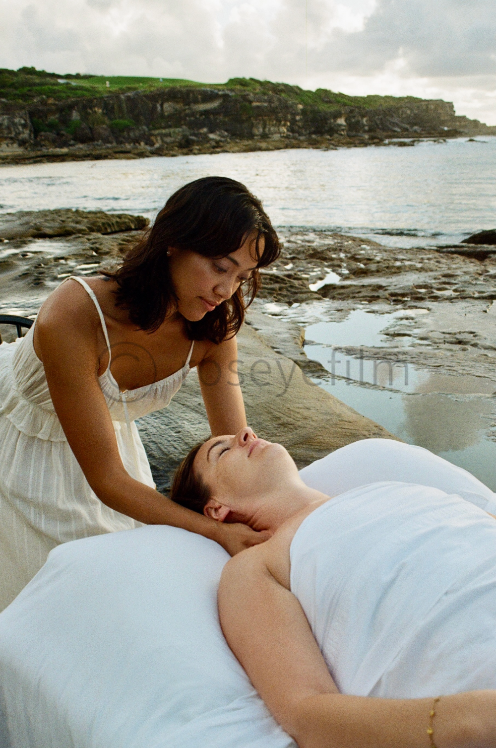 Two women, one lying on a bed and the other leaning over her, near a rocky shoreline with water and cliffs in the background.