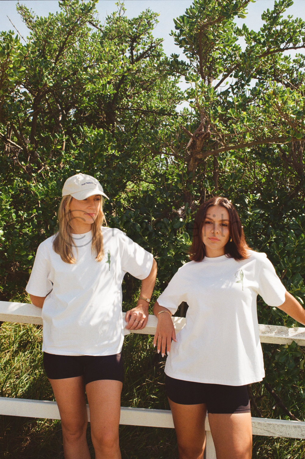 Two young women standing outdoors in front of a leafy green background, wearing white t-shirts and black shorts, with one wearing a white cap.