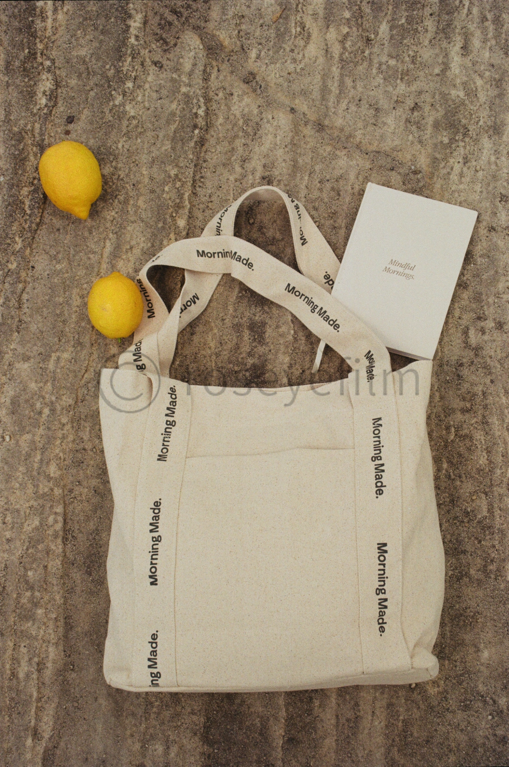 Two yellow lemons, a white tote bag with 'Morning Made.' printed on straps, and a small white card with 'Mindful Mornings.' are placed on a textured stone surface.