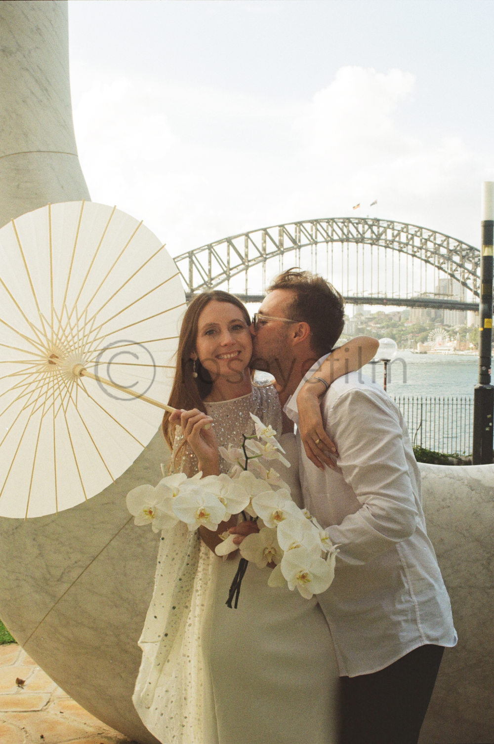 A newlywed couple celebrating in Sydney with the Harbour Bridge in the background. The woman is holding a white parasol and a bouquet of white orchids, and the man is kissing her cheek.
