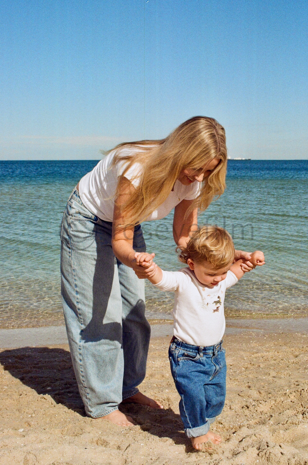 A woman helping a young child learn to walk on a sandy beach near the ocean, holding the child's hands and smiling.