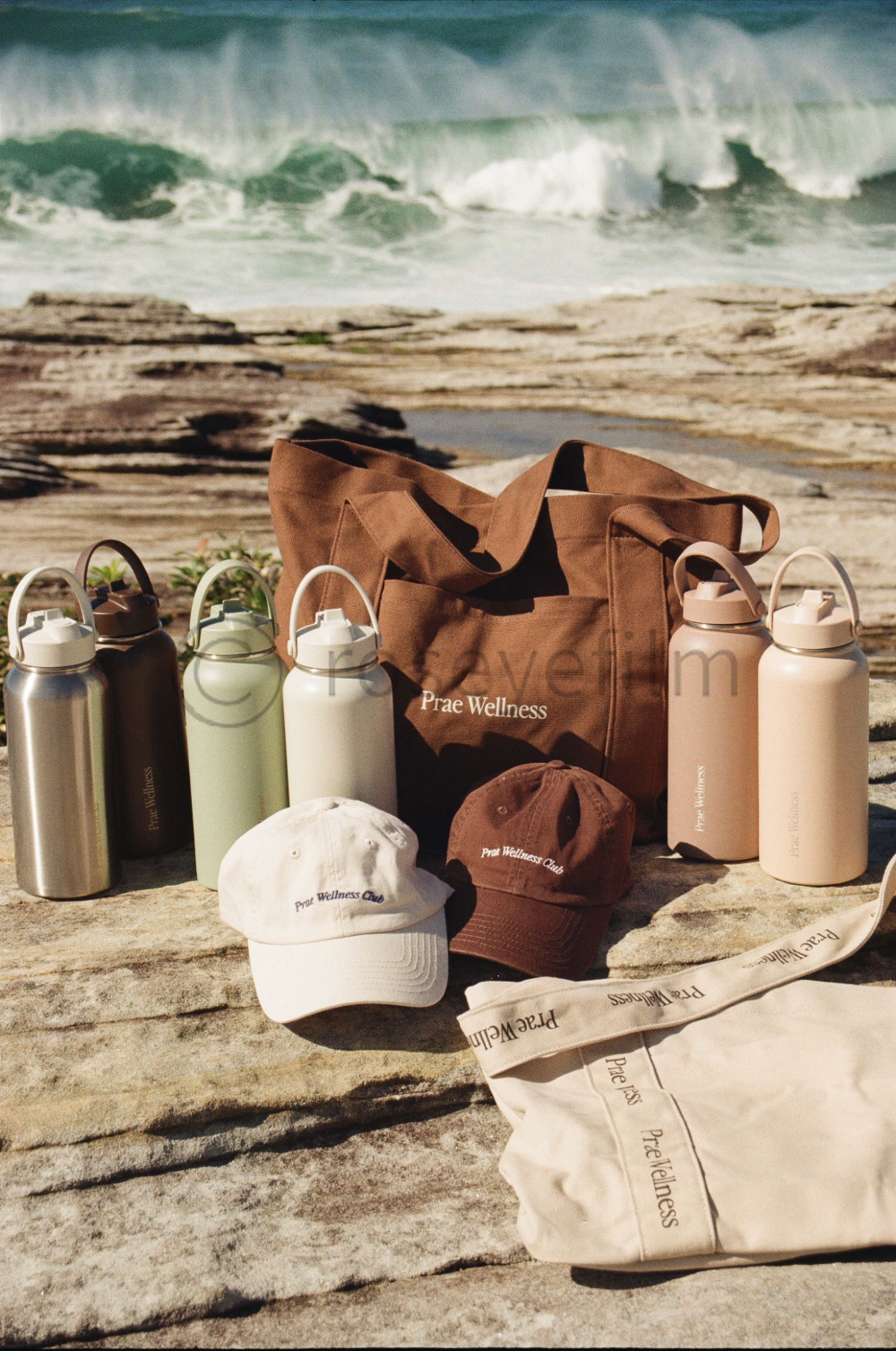 Collection of insulated water bottles, hats, tote bag, and a brown gym bag with 'Prae Wellness' branding on a rocky beach with ocean waves in the background.