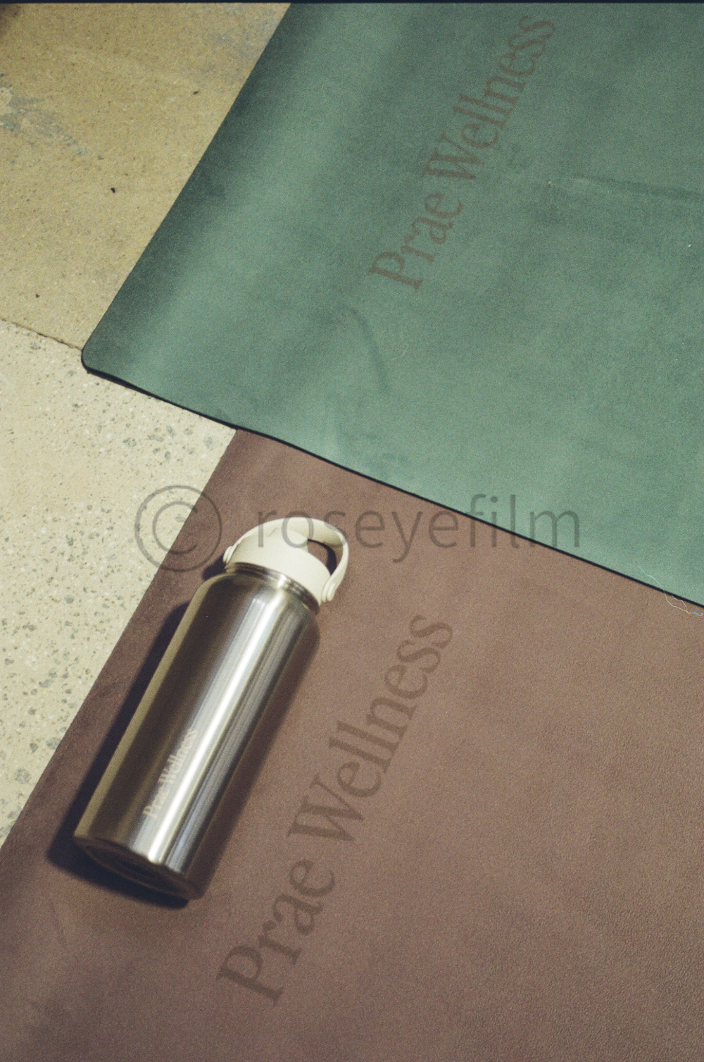 A stainless steel water bottle resting on a yoga mat, with a yoga mat and a piece of cloth with the words 'Prevale Wellness' printed on them, on a tiled floor.
