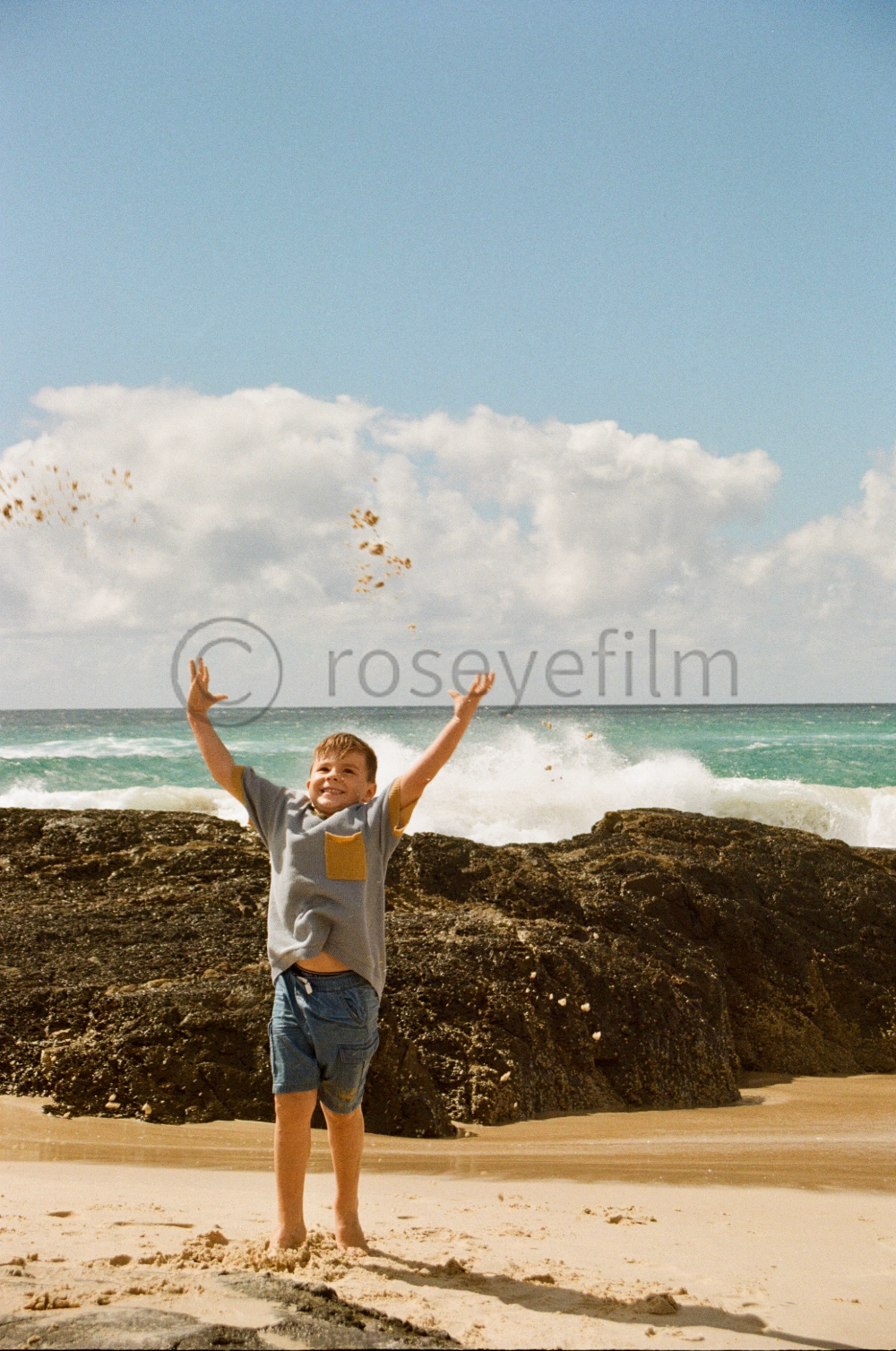 A young boy playing on a sandy beach near rocks with the ocean and cloudy sky in the background.