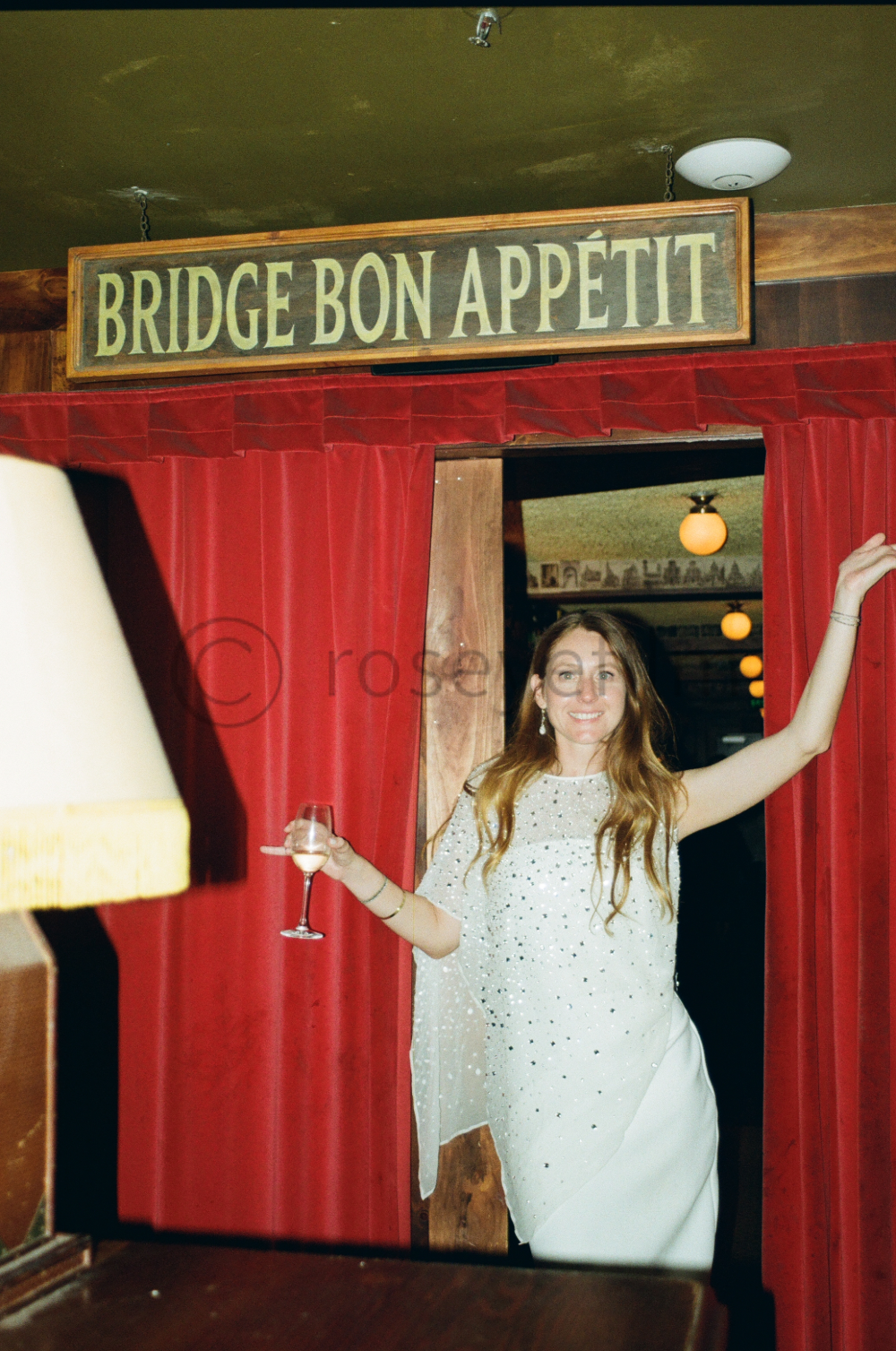 A woman with long, wavy hair holding a wine glass, standing in front of red curtains at an entrance with a sign that reads 'BRIDGE BON APPÉTIT' in a cozy, wood-paneled restaurant or bar.