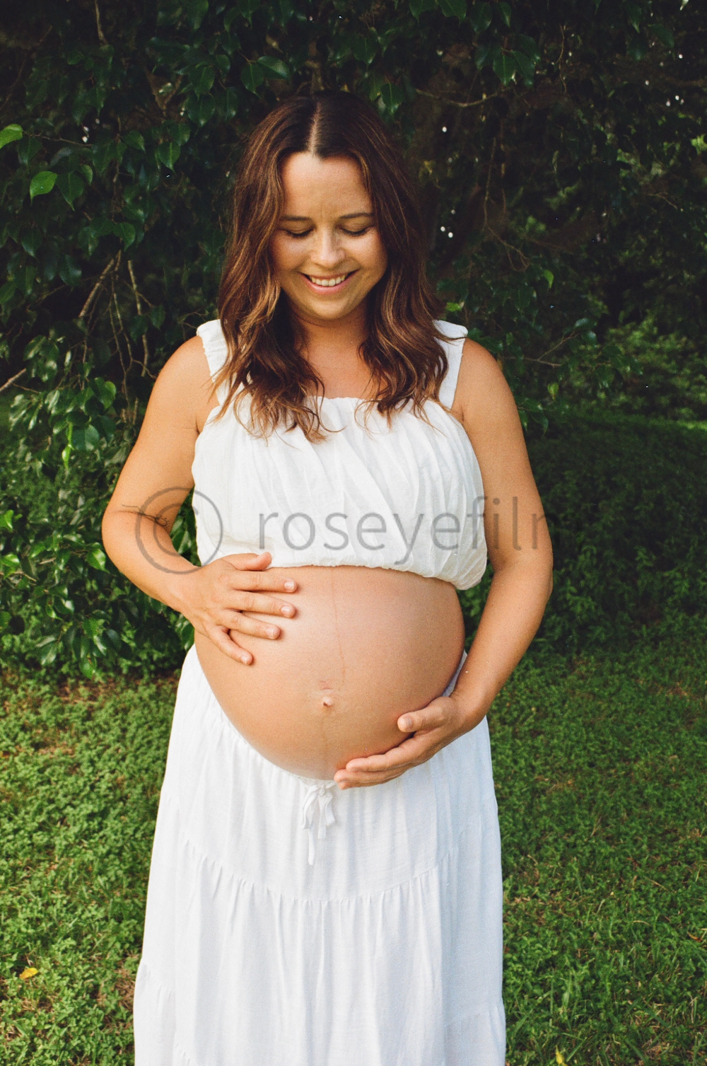 Pregnant woman in a white dress smiling and gently holding her baby bump outdoors with green foliage in the background.