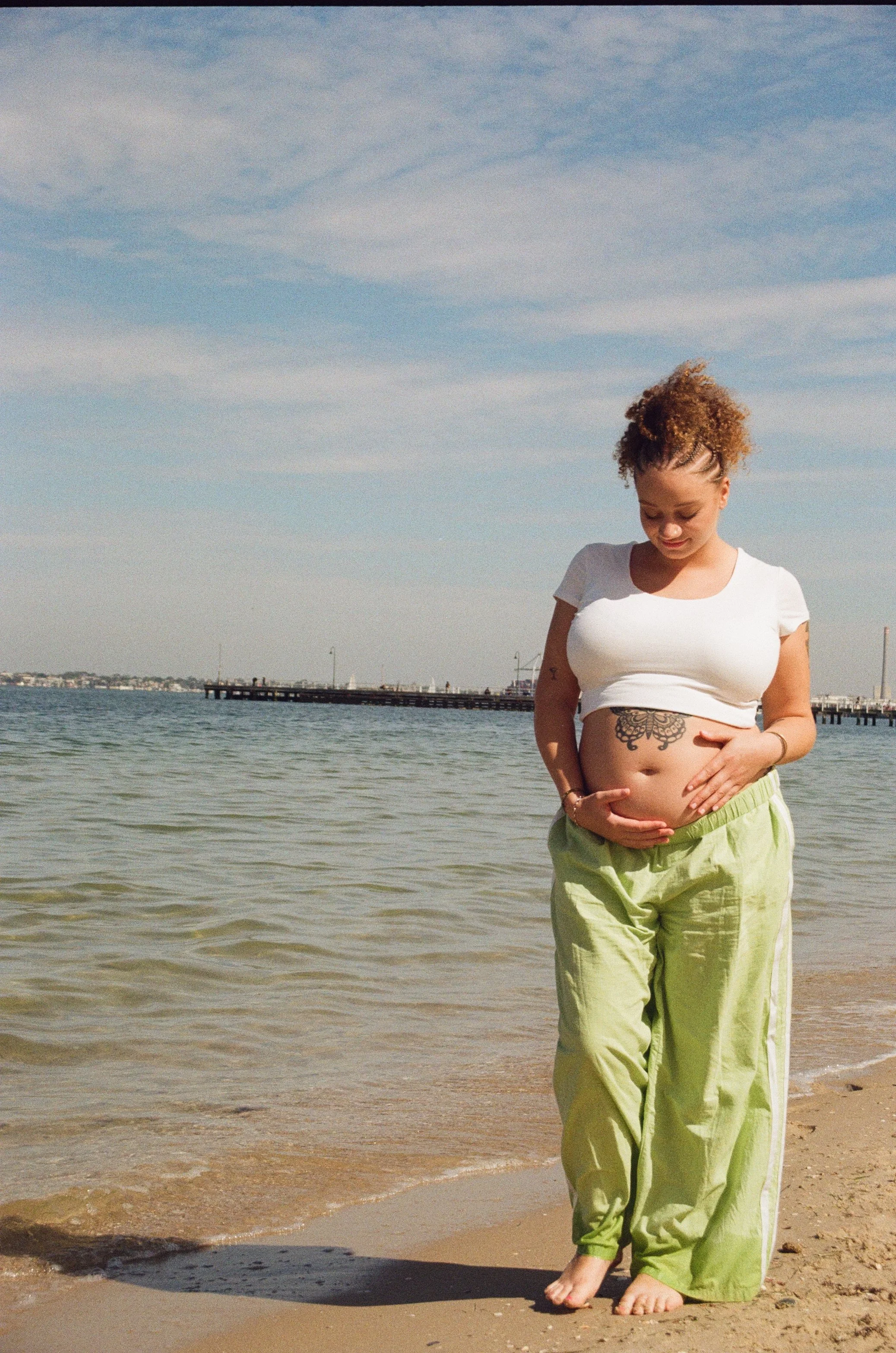 A woman standing at the beach with her hand on her pregnant belly, wearing light green pants and a white crop top, smiling while looking down, with water and a pier in the background under a blue sky with clouds.
