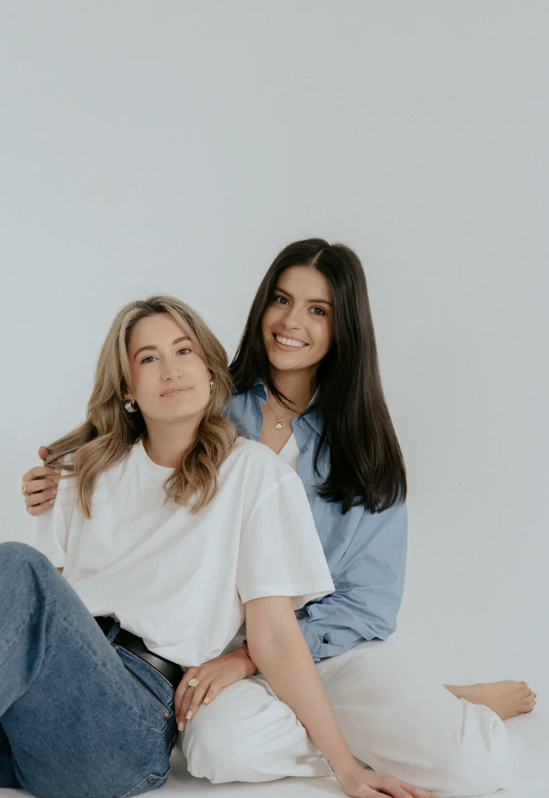 Two women sitting closely together against a plain white background, smiling and looking at the camera.