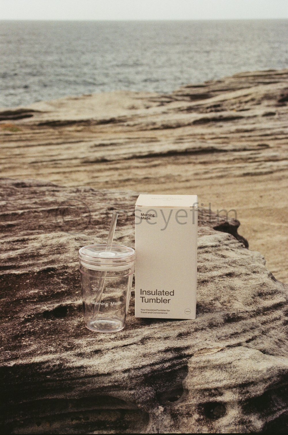 A clear insulated tumbler with a straw next to a white box labeled "Insulated Tumbler" on a large rocky surface near a beach and ocean in the background.