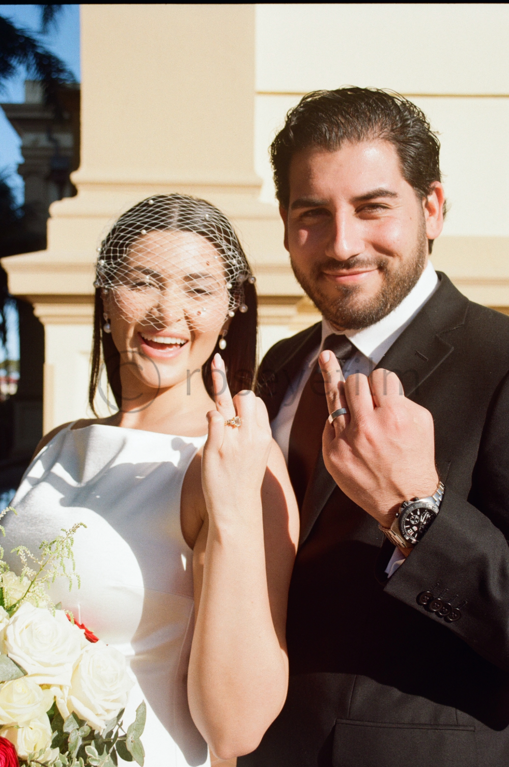 A newlywed couple in wedding attire, showing their wedding bands and making a victory sign with their hands, standing outdoors in front of a building.