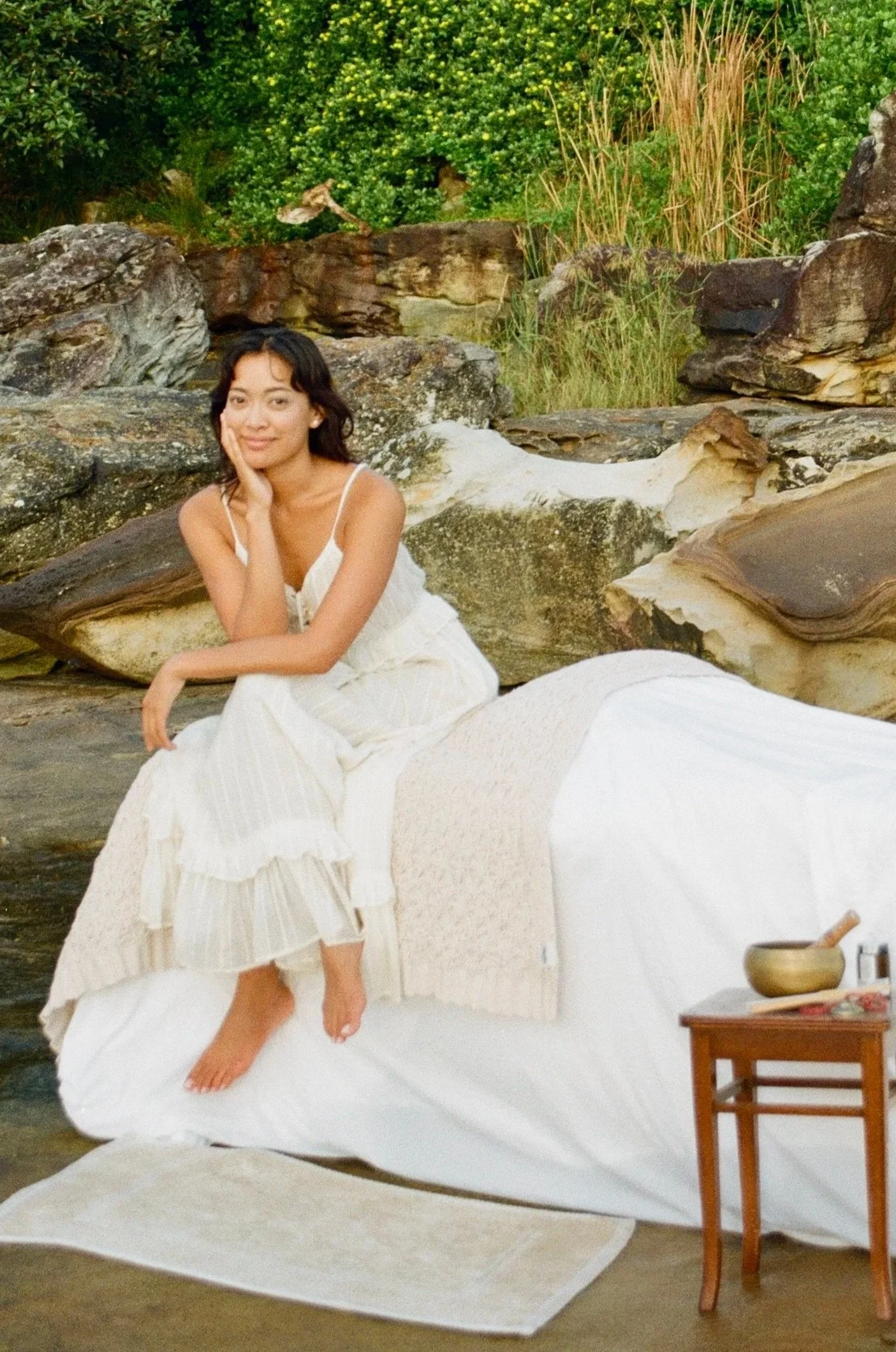 Woman sitting on a large, white, shell-shaped structure outdoors surrounded by rocks, greenery, and a wooden table with a bowl and other items.